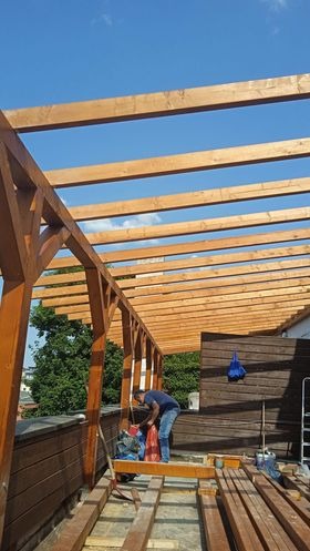Konstrukcja drewniana z belek na tle błękitnego nieba, widoczny pracownik w trakcie montażu. Elementy drewniane w ciepłym odcieniu brązu, częściowo zadaszone.