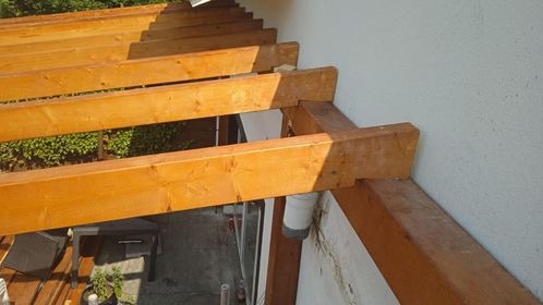 Drewniana konstrukcja z belek przy białej ścianie budynku, widoczny fragment rynny i zieleń w tle. Ujęcie z góry, podkreślające detale połączeń belek.