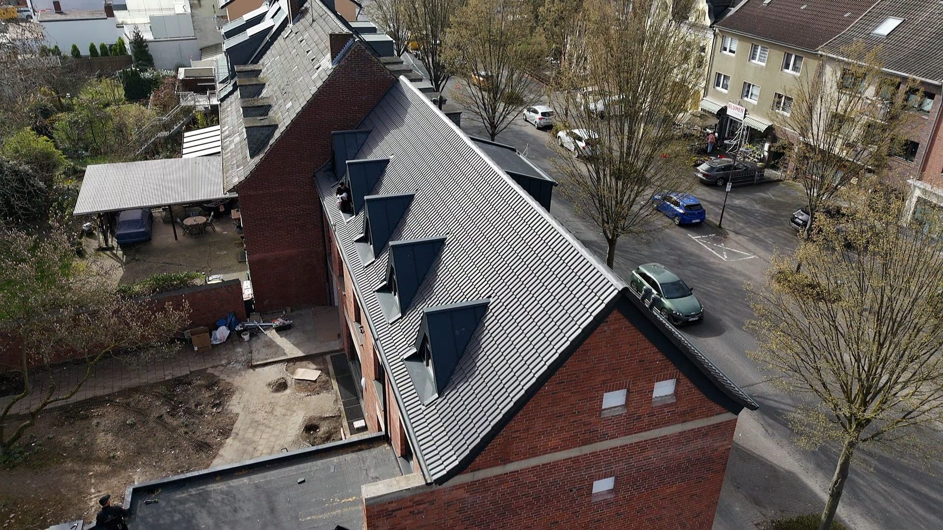 Czerwony budynek z nowym, szarym dachem z lukarnami, widziany z góry. W tle zabudowa miejska i drzewa bez liści. Widoczne detale wykończenia dachu.