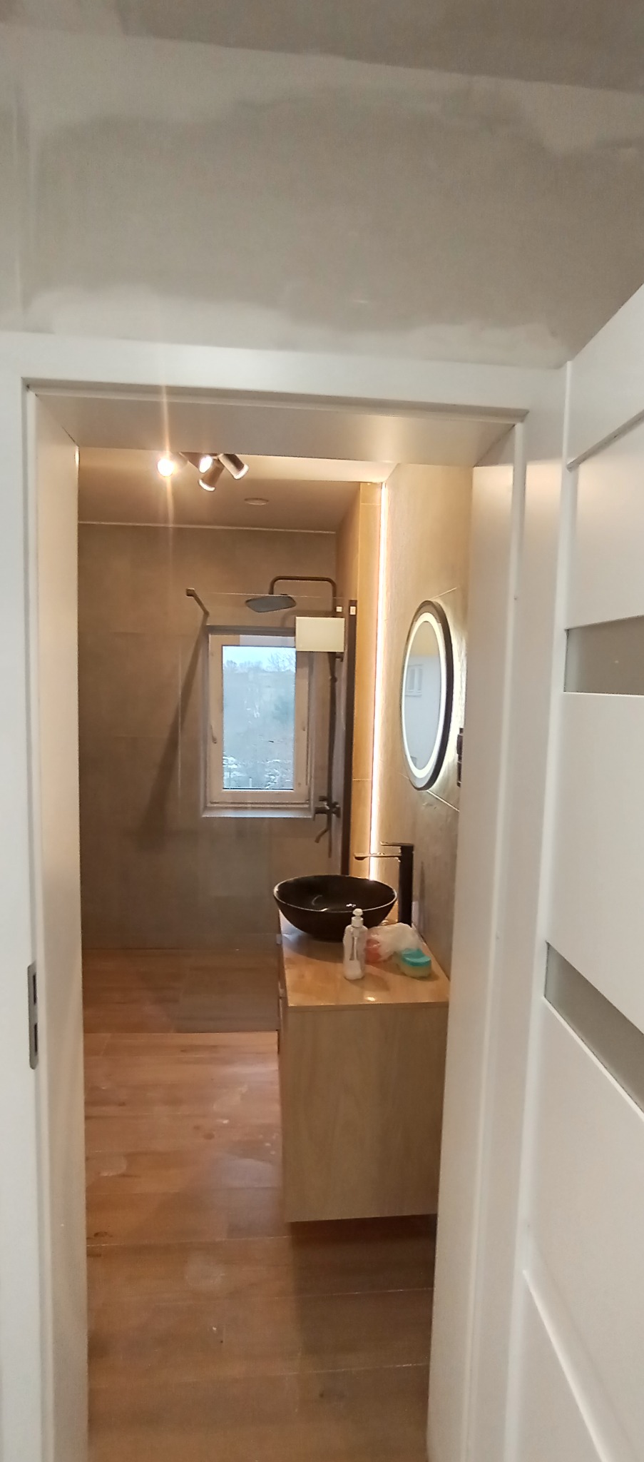 Nowoczesna łazienka po remoncie: drewniana podłoga, umywalka w kształcie misy, okrągłe lustro z podświetleniem LED i kabina prysznicowa z deszczownicą.