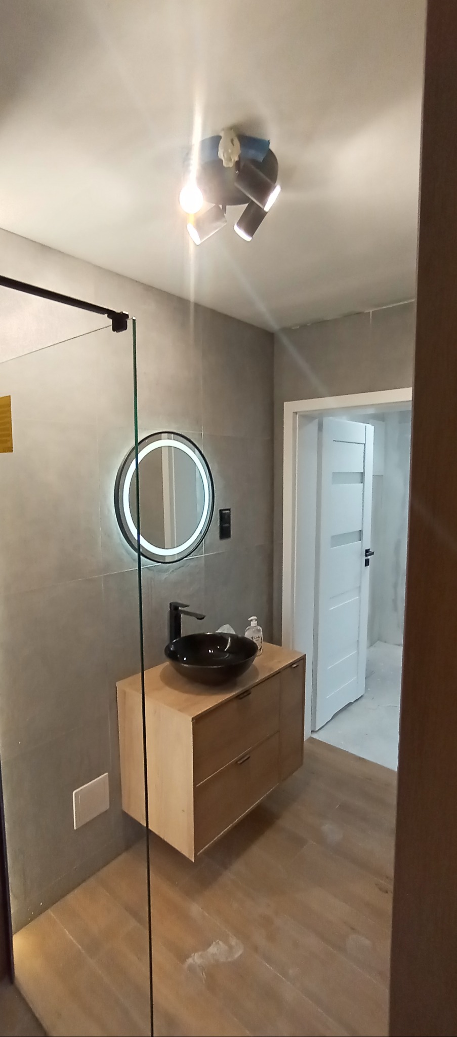 Nowoczesna łazienka z drewnianą szafką pod umywalką, okrągłym lustrem z podświetleniem LED i szklaną kabiną prysznicową. Szare płytki na ścianach i drewniana podłoga.