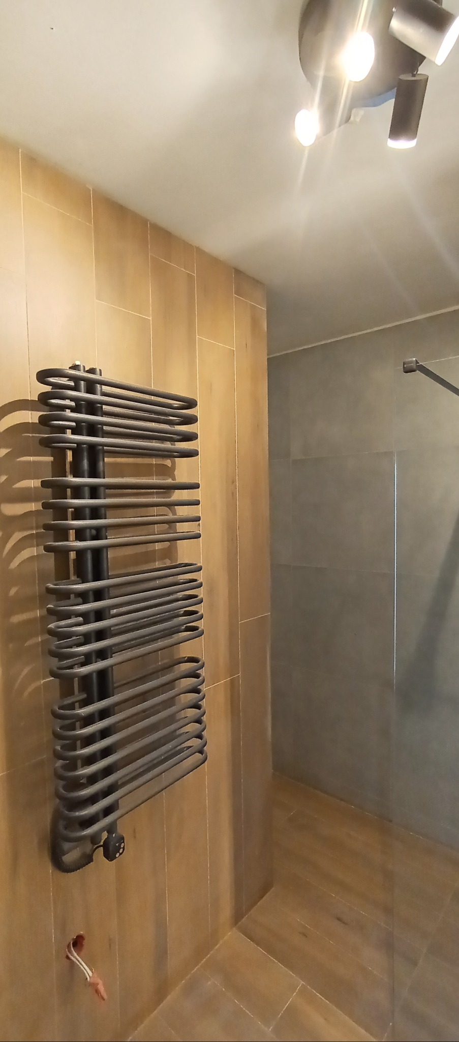 Zamontowany, grafitowy grzejnik łazienkowy na ścianie z płytek imitujących drewno. W tle widoczny prysznic i oświetlenie sufitowe.