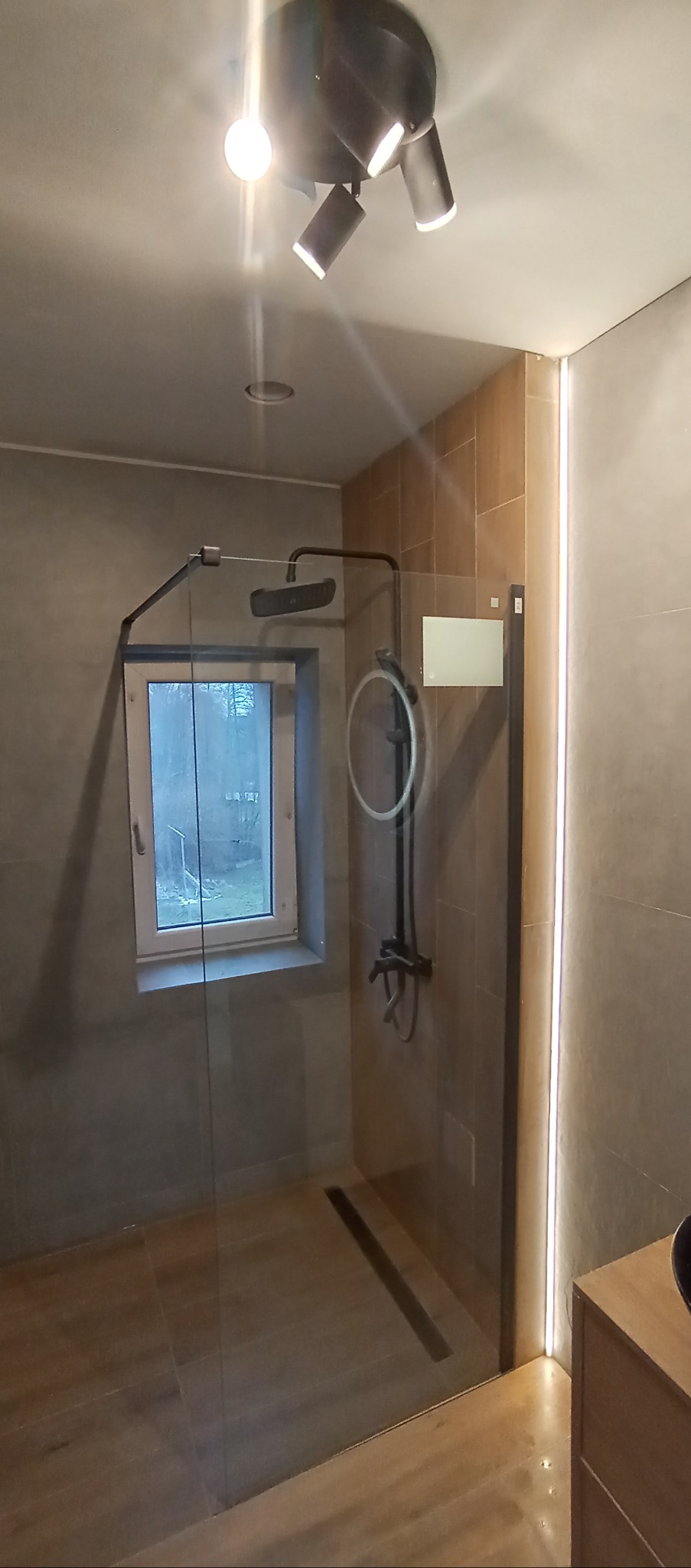 Nowoczesna kabina prysznicowa z czarną armaturą i odpływem liniowym, oświetlona punktowo, z widokiem na okno. Minimalistyczny design, jasne płytki i drewnopodobna podłoga.