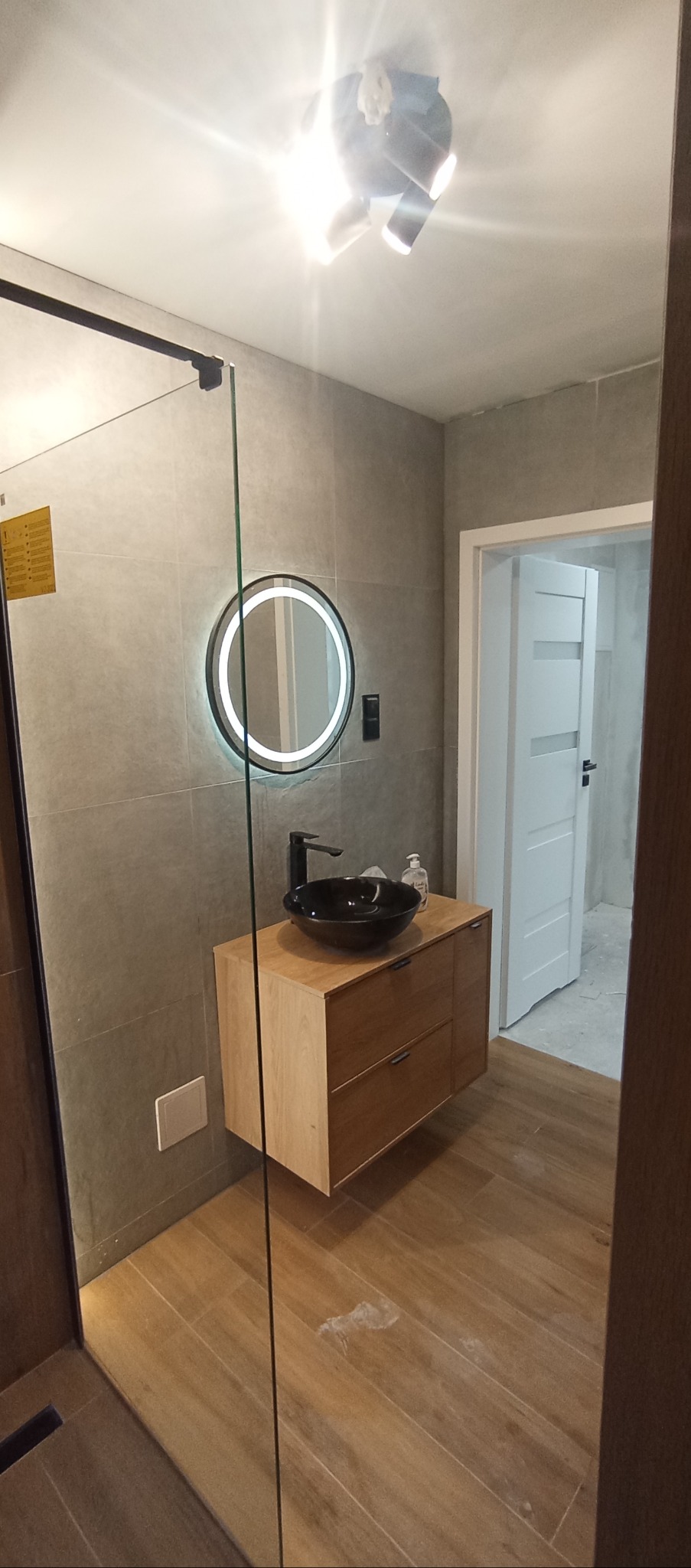 Nowoczesna łazienka z drewnianą szafką pod umywalkę w kształcie misy, okrągłym lustrem z podświetleniem LED i szklaną kabiną prysznicową.