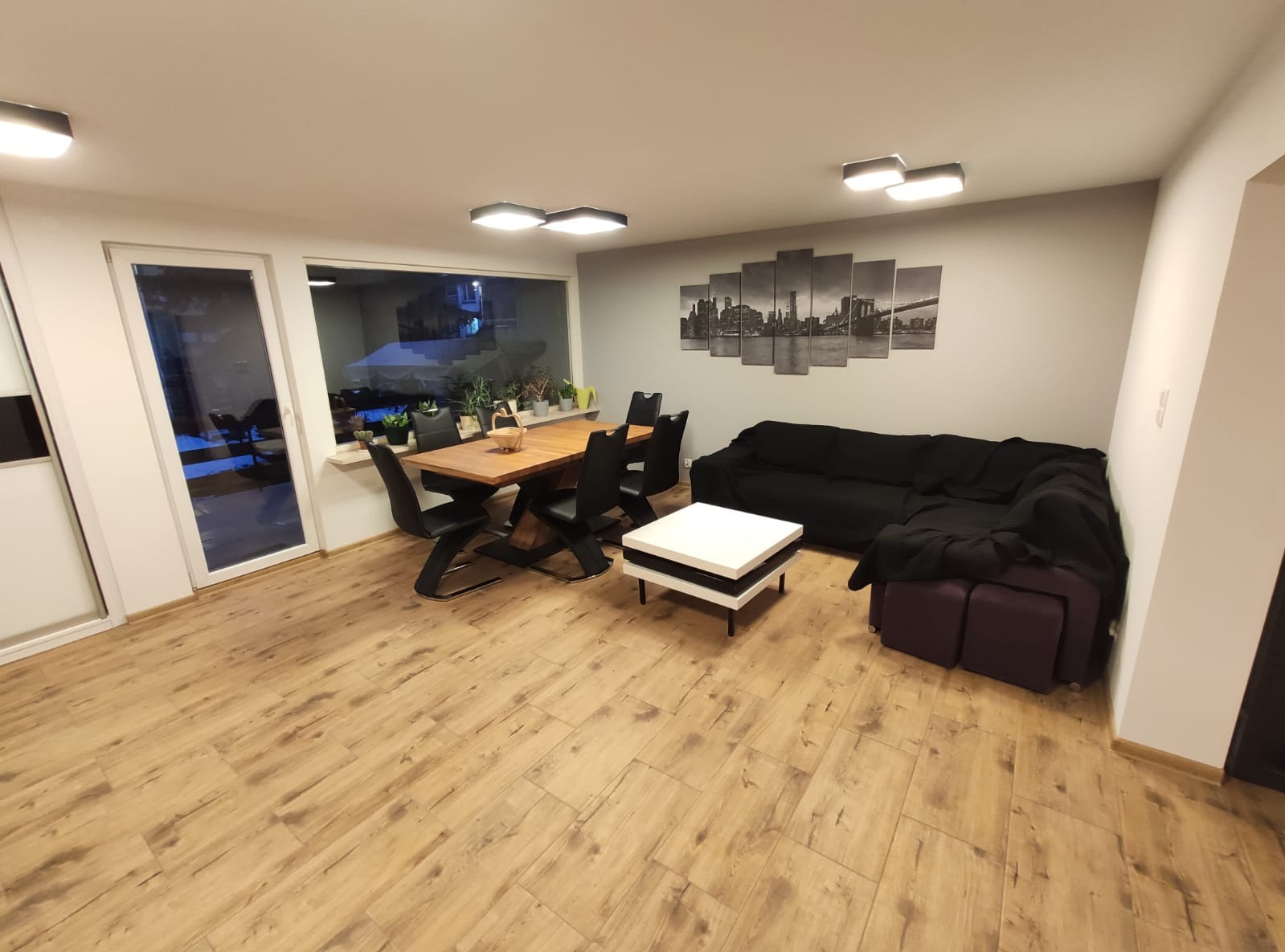 Jasne wnętrze z drewnianą podłogą, stołem z krzesłami i czarną sofą. Na ścianie obraz z motywem miejskim. Nowoczesne oświetlenie sufitowe.