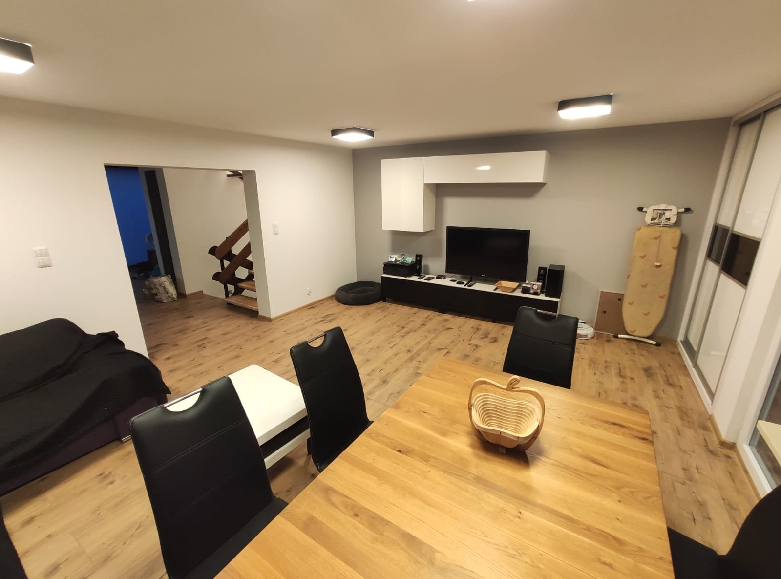 Jasne wnętrze salonu z drewnianą podłogą i stołem. W tle telewizor, schody i deska do prasowania. Minimalistyczny design z elementami dekoracyjnymi.