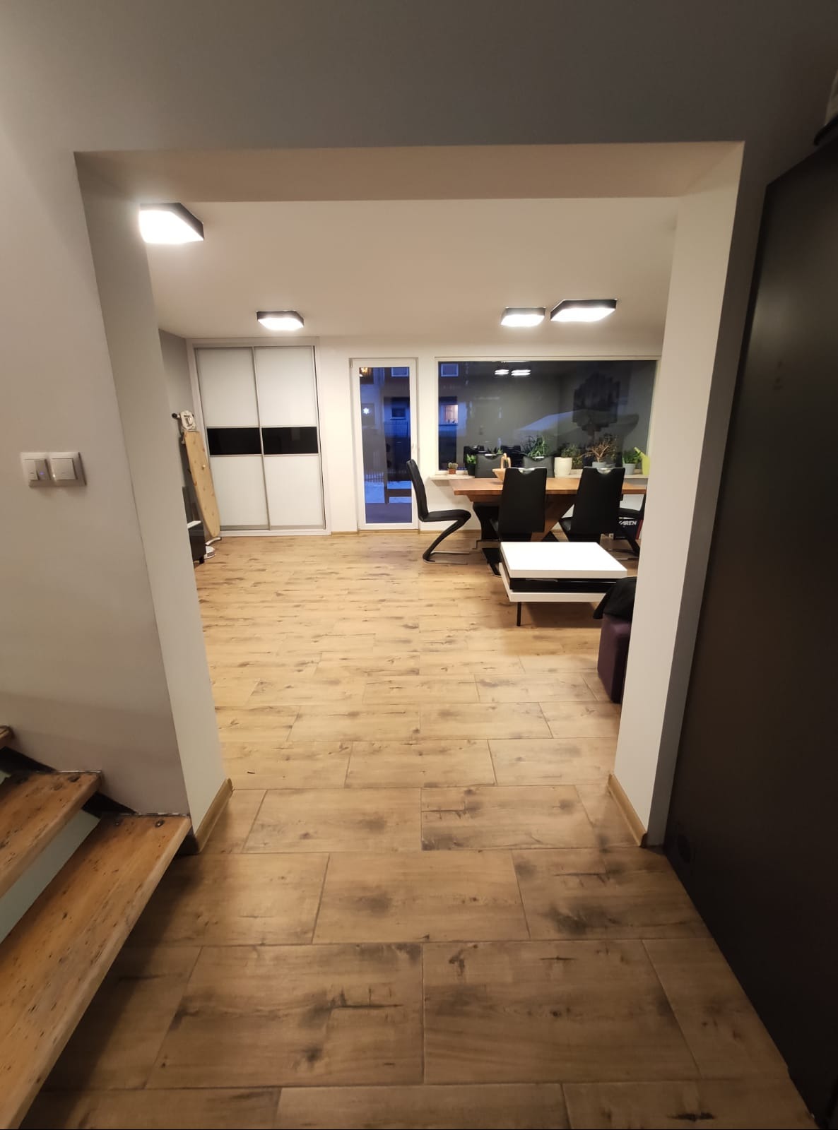 Salon z drewnianą podłogą, widok z korytarza. Jasne wnętrze z dużym oknem, minimalistyczne meble. Podłoga ułożona w deski, ciepłe oświetlenie.