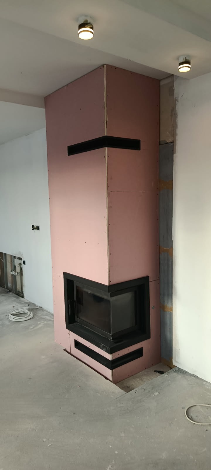 Wnętrze w trakcie prac wykończeniowych: obudowa kominka z płyt kartonowo-gipsowych w kolorze różowym, czarny wkład kominkowy, surowy beton na podłodze, widoczne instalacje.