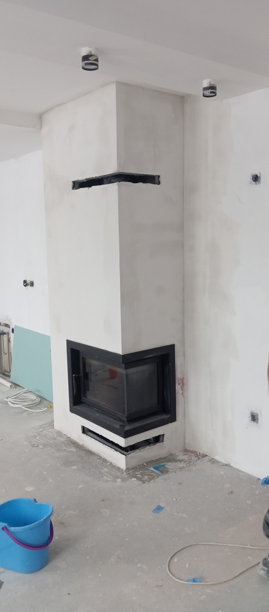 Narożny kominek z czarną obudową w surowym, białym wykończeniu. Widoczne niedoskonałości szpachlowania i otwory wentylacyjne. W tle ściany w trakcie prac wykończeniowych.