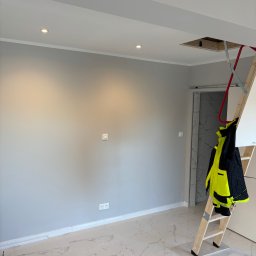 M.W.INVEST - Świeżo pomalowane szare wnętrze z marmurową podłogą, widoczne gniazdka, włącznik, drabina oparta o ścianę i otwór w suficie.