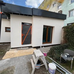 M.W.INVEST - Otynkowany budynek z oknami i zabezpieczonymi drzwiami, widoczne puszki z farbą i drabina malarska. Elewacja w jasnym odcieniu, tło stanowi budynek mieszkalny.