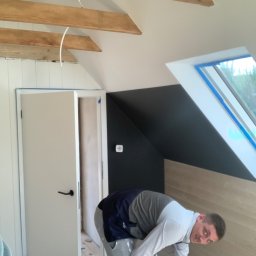 M.W.INVEST - Prace wykończeniowe na poddaszu: mężczyzna szlifuje drewnianą podłogę w pomieszczeniu ze skosami, widoczne drewniane belki i okno dachowe.