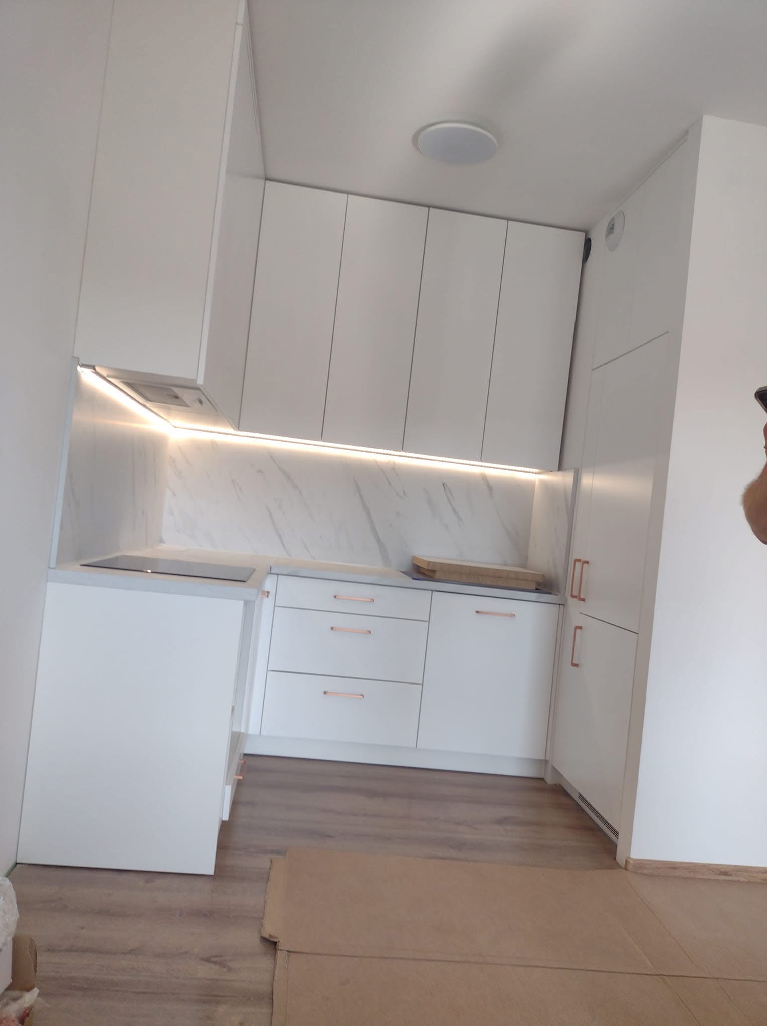 Biała kuchnia na wymiar z marmurowym blatem i oświetleniem LED podszafkowym. Miedziane uchwyty dodają elegancji. Widoczne panele podłogowe.