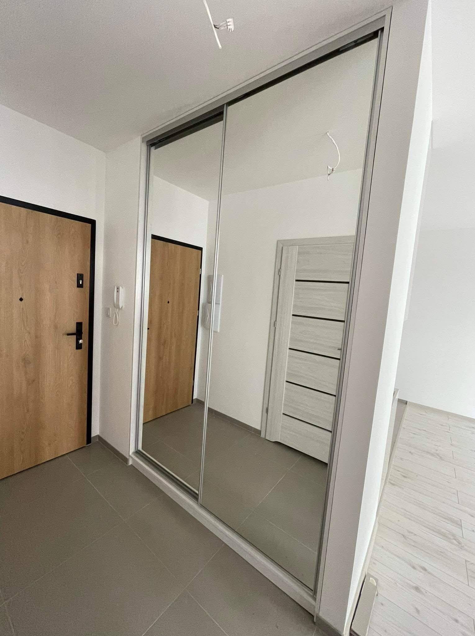 Nowoczesna szafa wnękowa z lustrzanymi drzwiami przesuwnymi, wkomponowana w jasne wnętrze przedpokoju z szarą podłogą i dwiema parami drzwi.