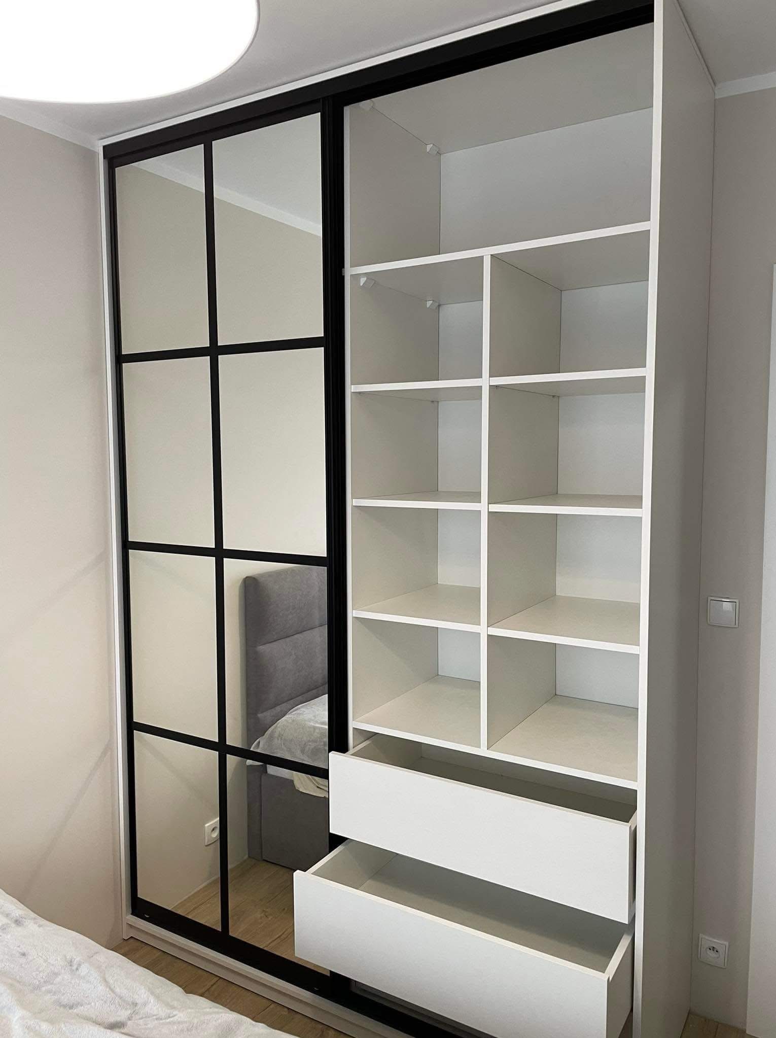 Biała szafa wnękowa z przesuwnymi drzwiami z lustrem i czarnymi szprosami. Część szafy otwarta z półkami i szufladami. Widoczny fragment łóżka.