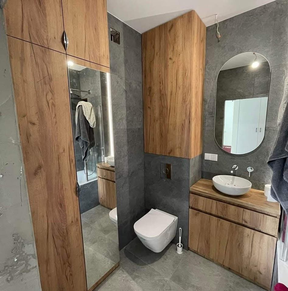 Nowoczesna łazienka z drewnianymi szafkami, owalnym lustrem i podwieszaną toaletą. Szare płytki na ścianach i podłodze. Szafka z lustrem po lewej stronie.