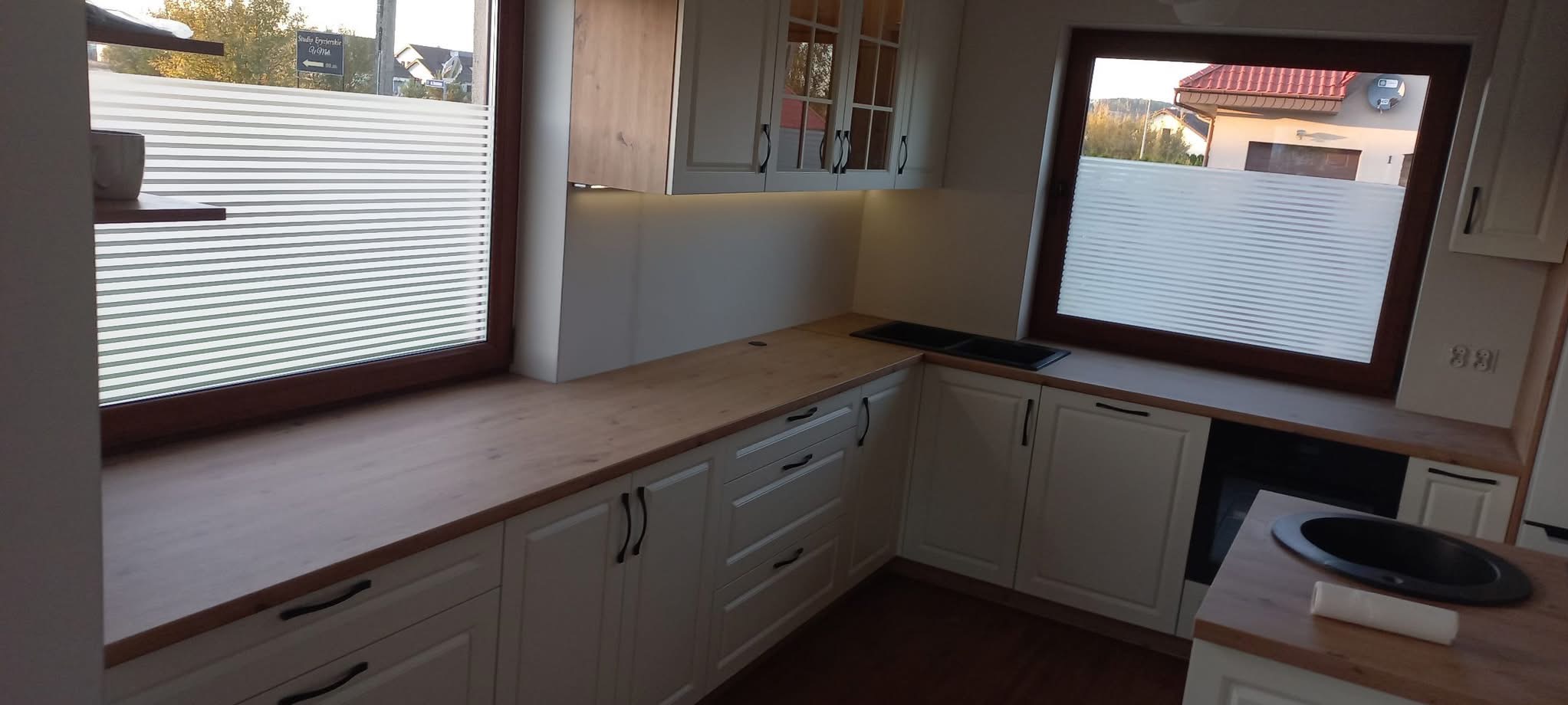 Jasna kuchnia z drewnianym blatem i białymi frontami szafek, z oknami przesłoniętymi żaluzjami plisowanymi. Widoczny zlew i fragment zabudowy piekarnika.
