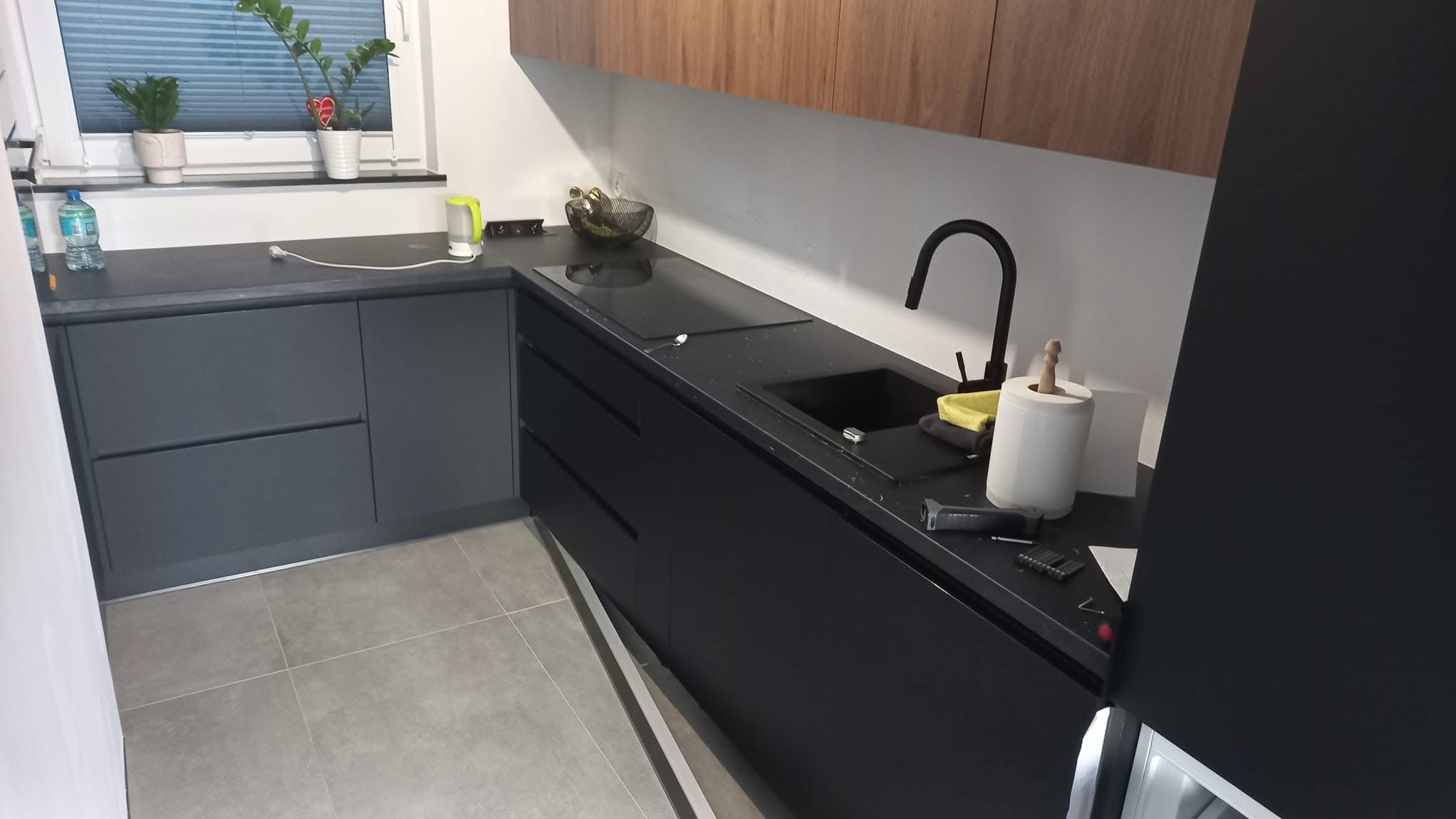 Nowoczesna kuchnia z czarnym blatem i frontami, widoczne narzędzia, zlewozmywak i płyta indukcyjna. U góry szafki z drewnianym wykończeniem. Na parapecie rośliny doniczkowe.