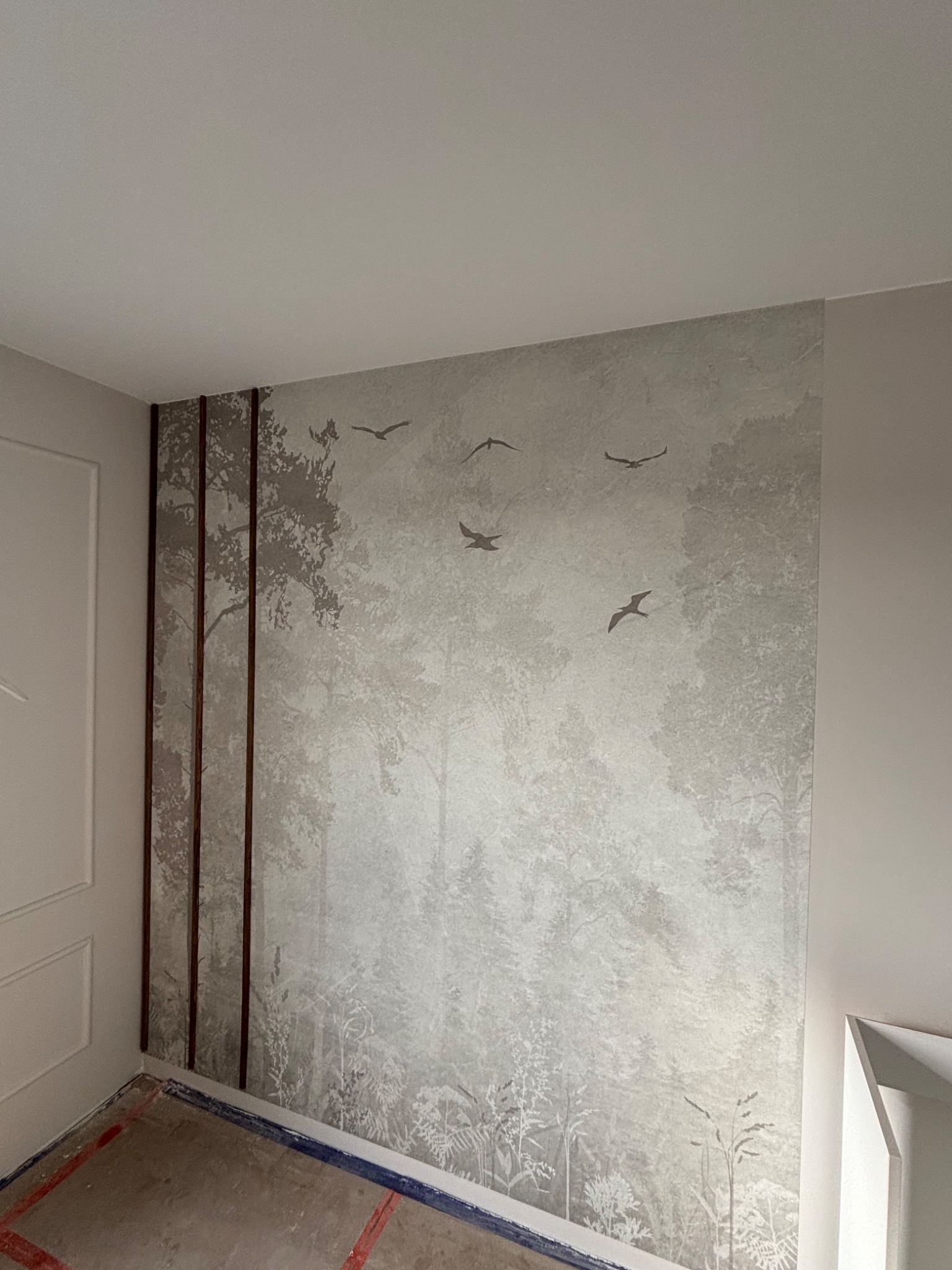 Ściana z tapetą w leśny motyw z sylwetkami ptaków, ozdobiona pionowymi listwami. Podłoga przygotowana do dalszych prac wykończeniowych.