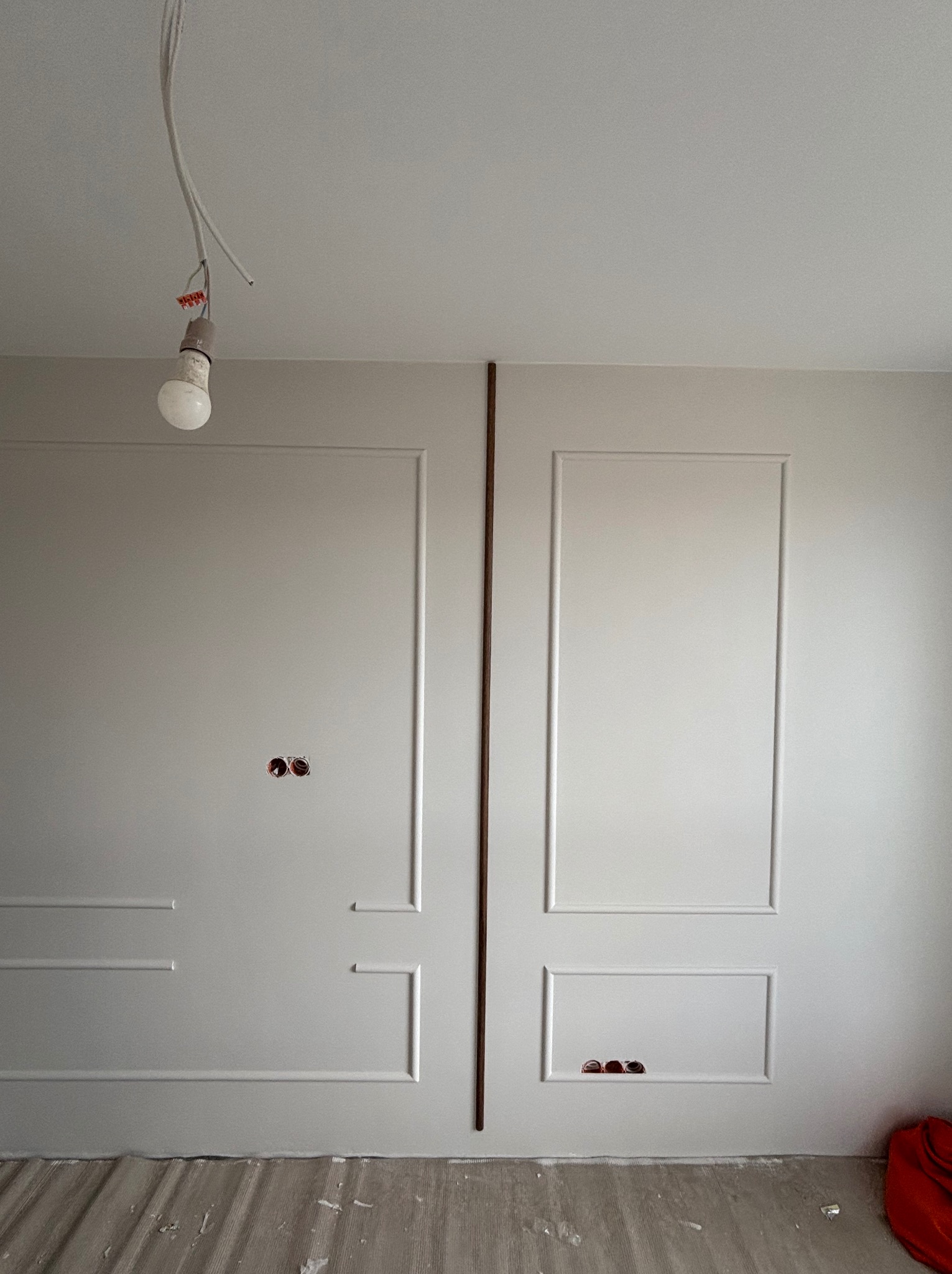 Ściana z dekoracyjnymi panelami w trakcie montażu, widoczne gniazdka elektryczne i prowizoryczne oświetlenie. Podłoga zabezpieczona folią malarską.