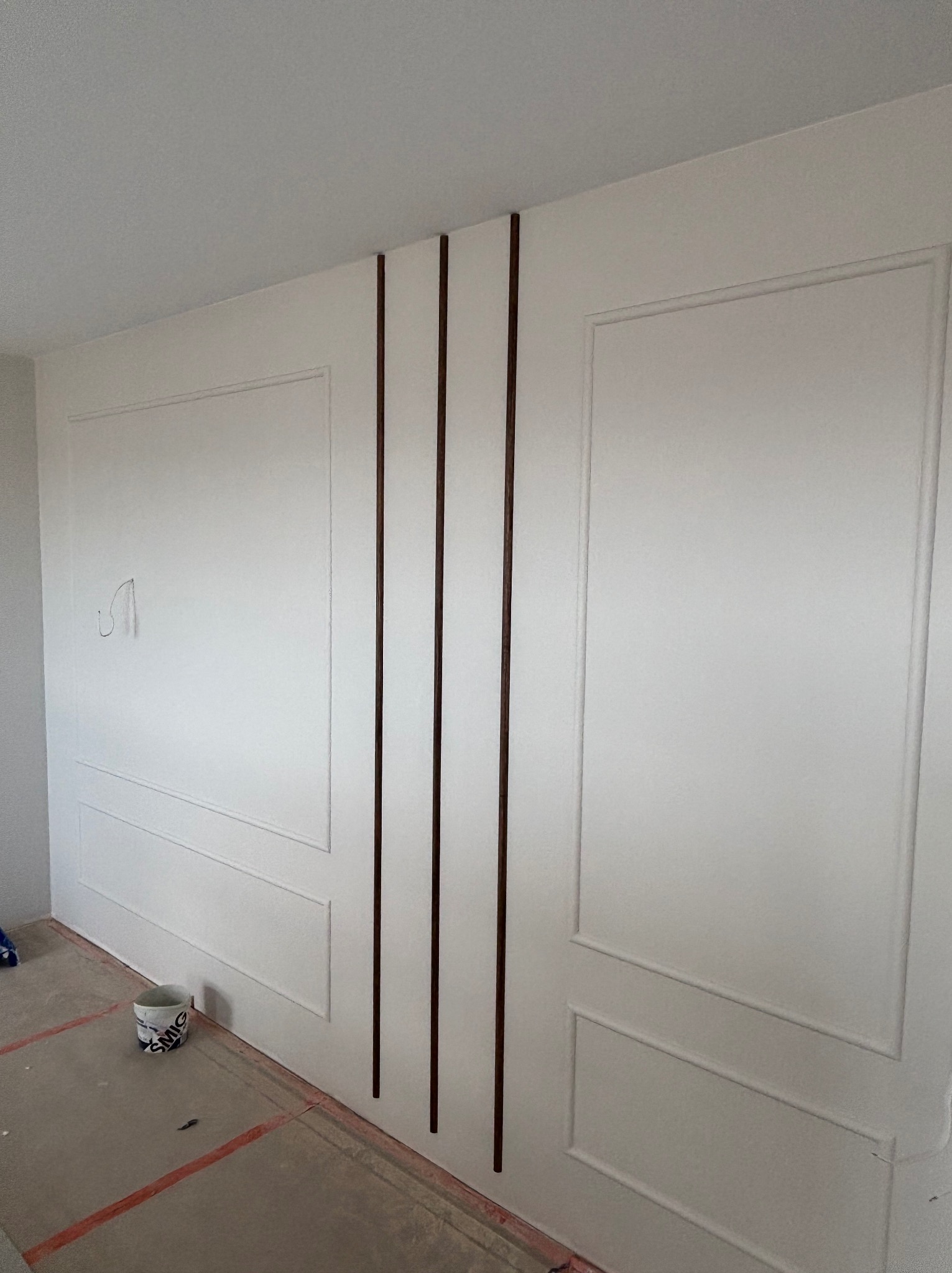 Biała ściana z dekoracyjnymi panelami i brązowymi listwami. Widoczny fragment podłogi z zabezpieczeniem malarskim i wiadro z farbą. Detal wykończenia wnętrza.