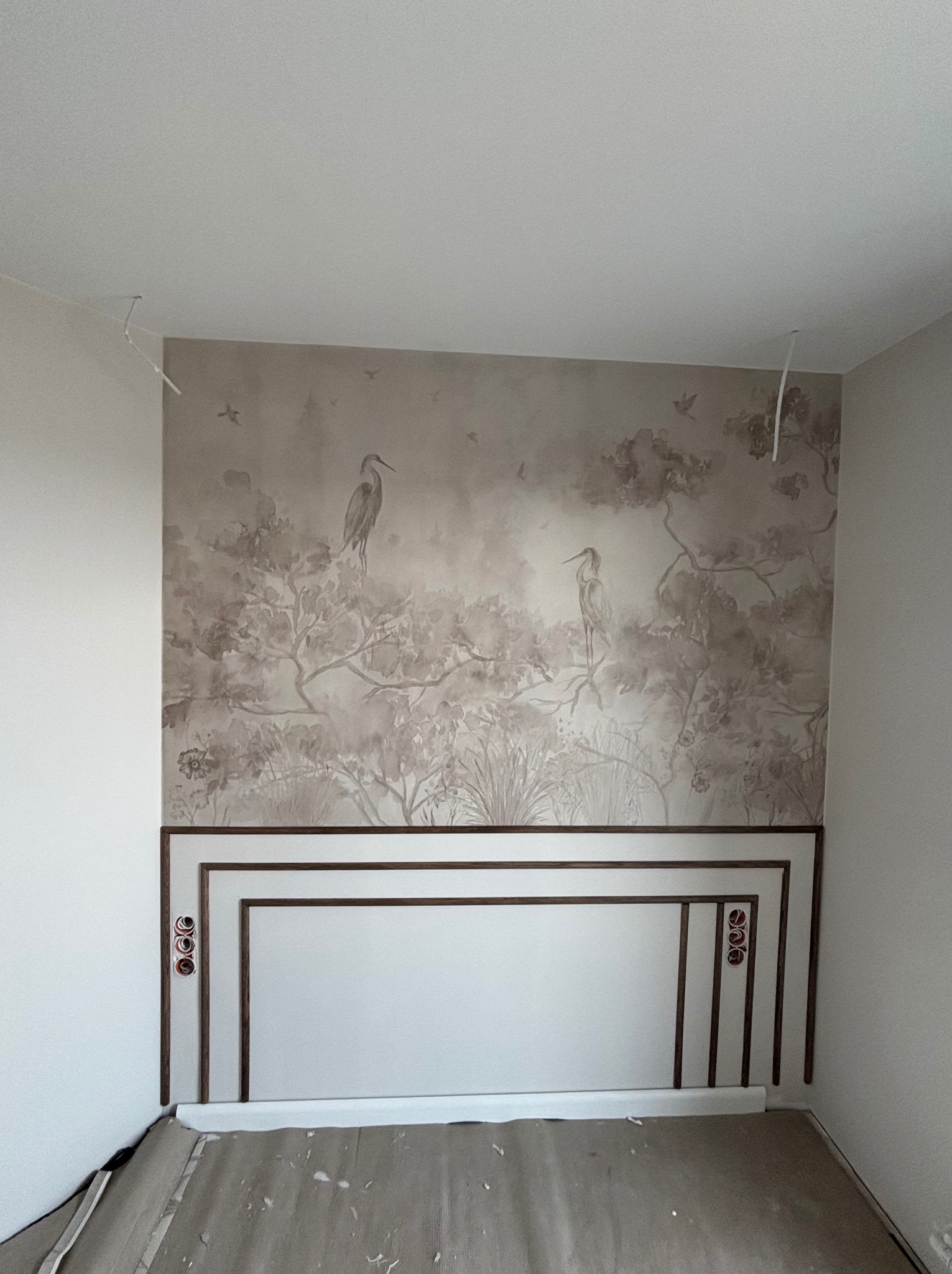 Ściana z dekoracyjnym panelem i tapetą w ptaki, widoczne gniazdka elektryczne. Podłoga częściowo zakryta folią ochronną. Wnętrze w trakcie wykończenia.