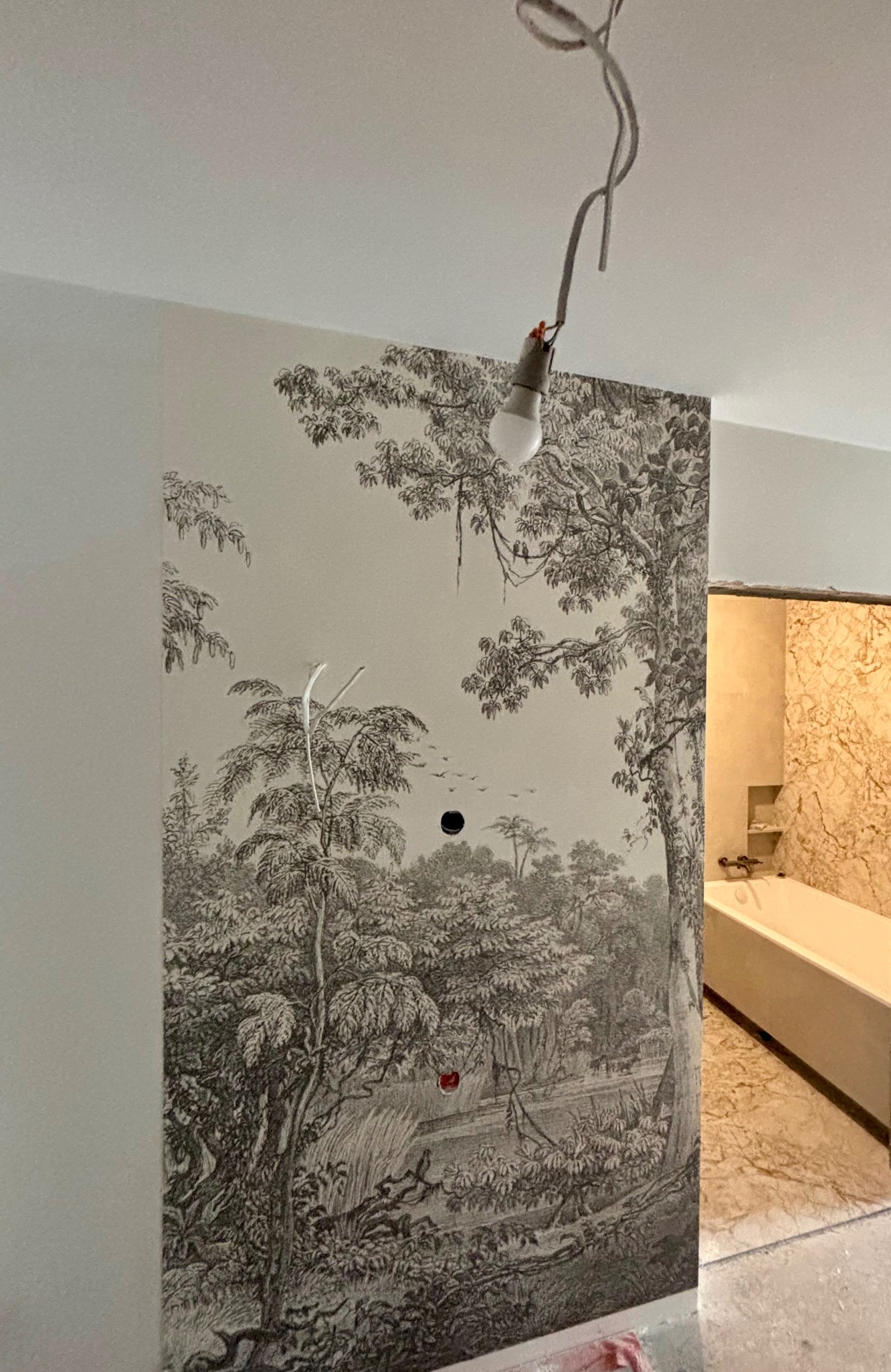 Tapeta z motywem leśnym na ścianie łazienki w trakcie montażu, widoczne przewody oświetleniowe i fragment wanny. Dekoracja wnętrza w stylu klasycznym.