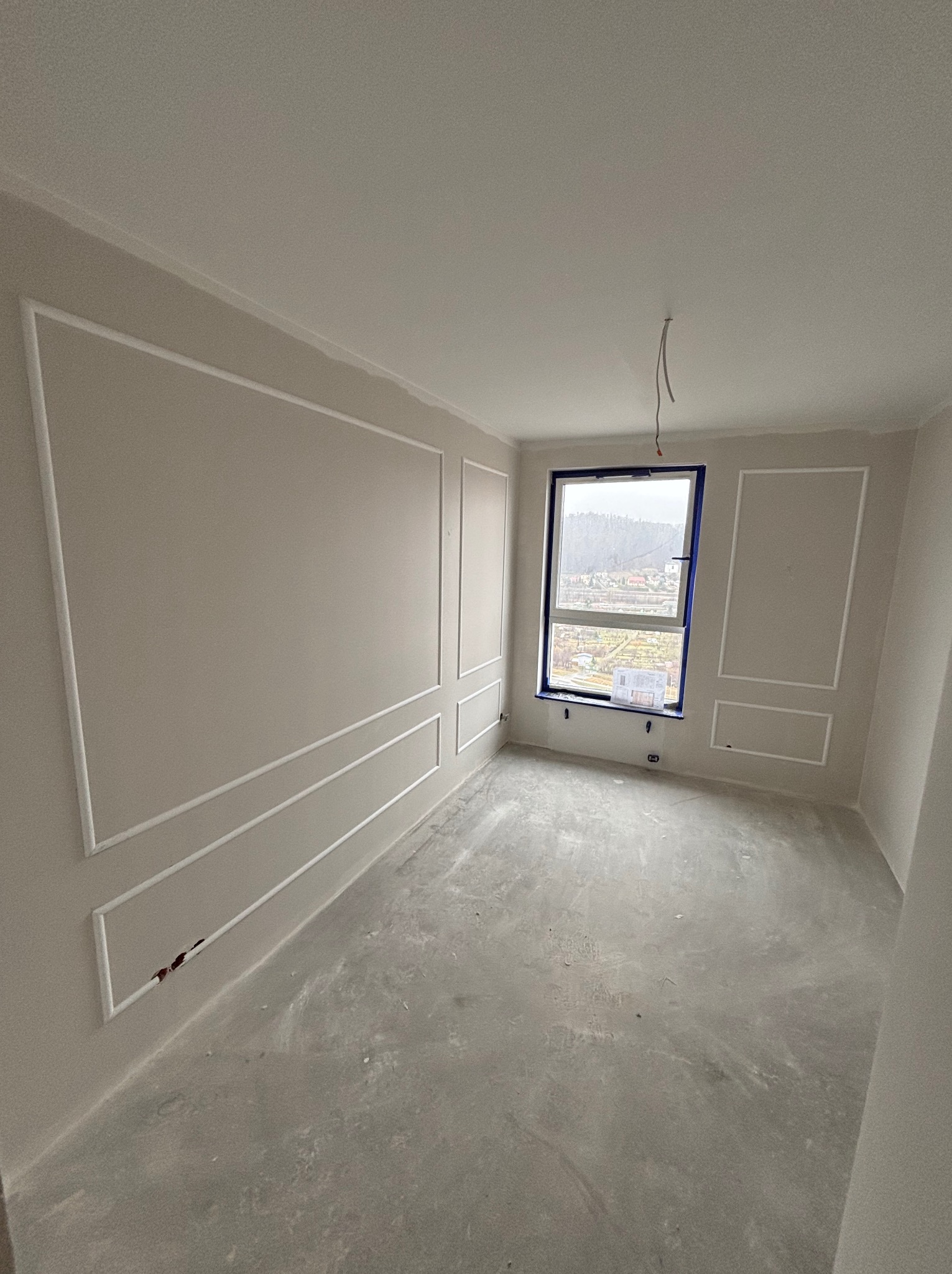 Jasne wnętrze pokoju z dekoracyjnymi listwami ściennymi i oknem z widokiem na miasto. Surowa podłoga, przygotowana do dalszych prac wykończeniowych.