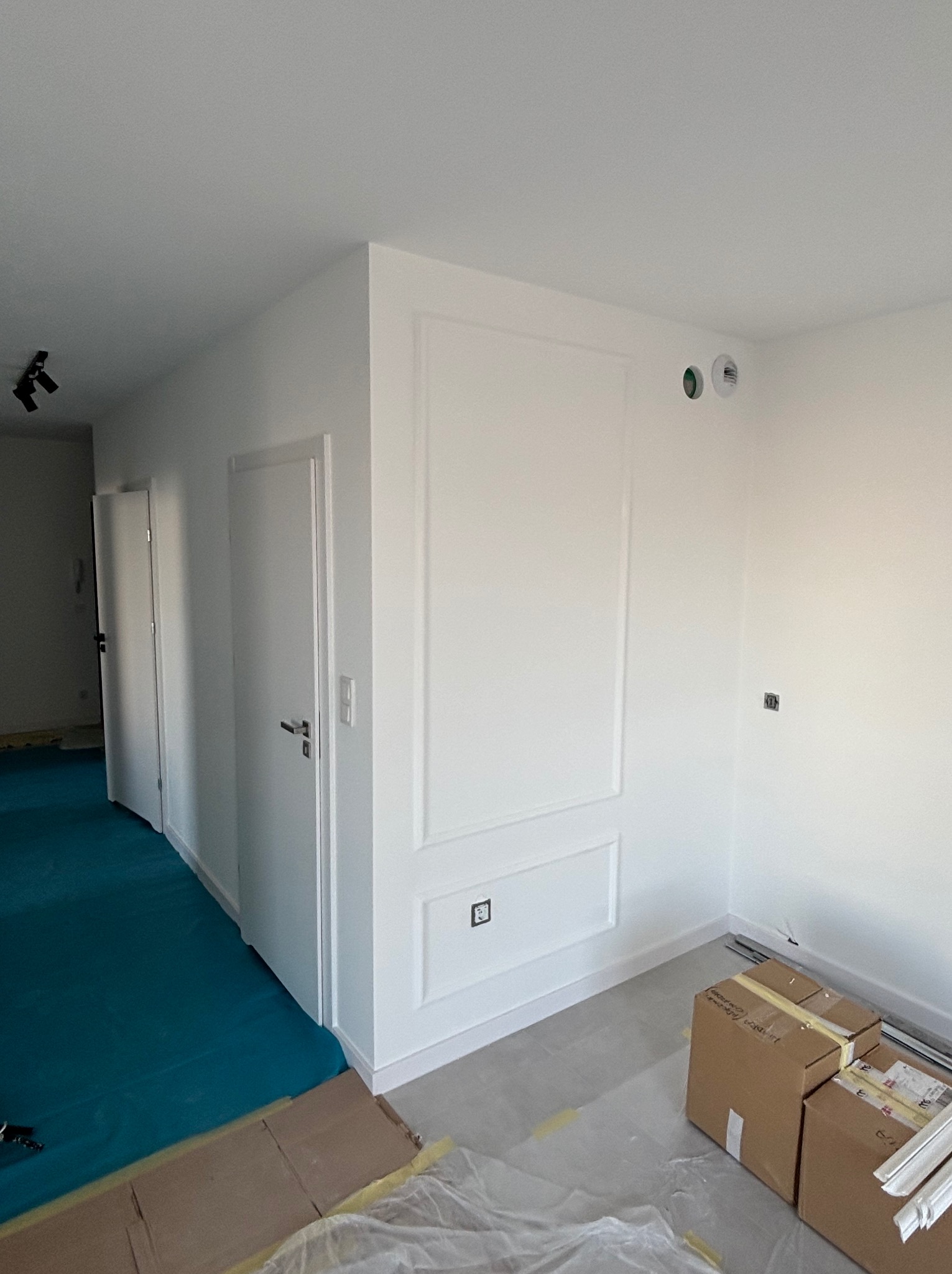 Białe, odświeżone wnętrze z dekoracyjną sztukaterią na ścianie, widoczne drzwi i zabezpieczona podłoga. Kartony i listwy przypodłogowe sugerują prace wykończeniowe.