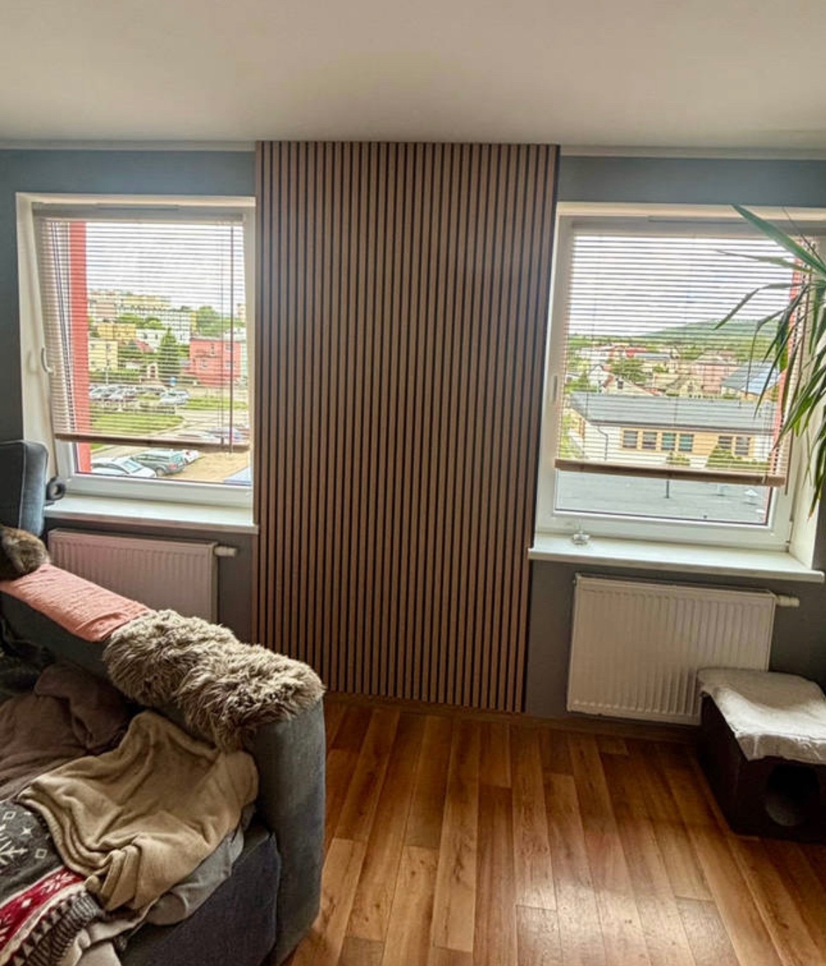 Panel ścienny z drewnianych listew w salonie, widok na okno z żaluzjami i miasto w tle. Parkiet na podłodze, fragment sofy i rośliny doniczkowej.