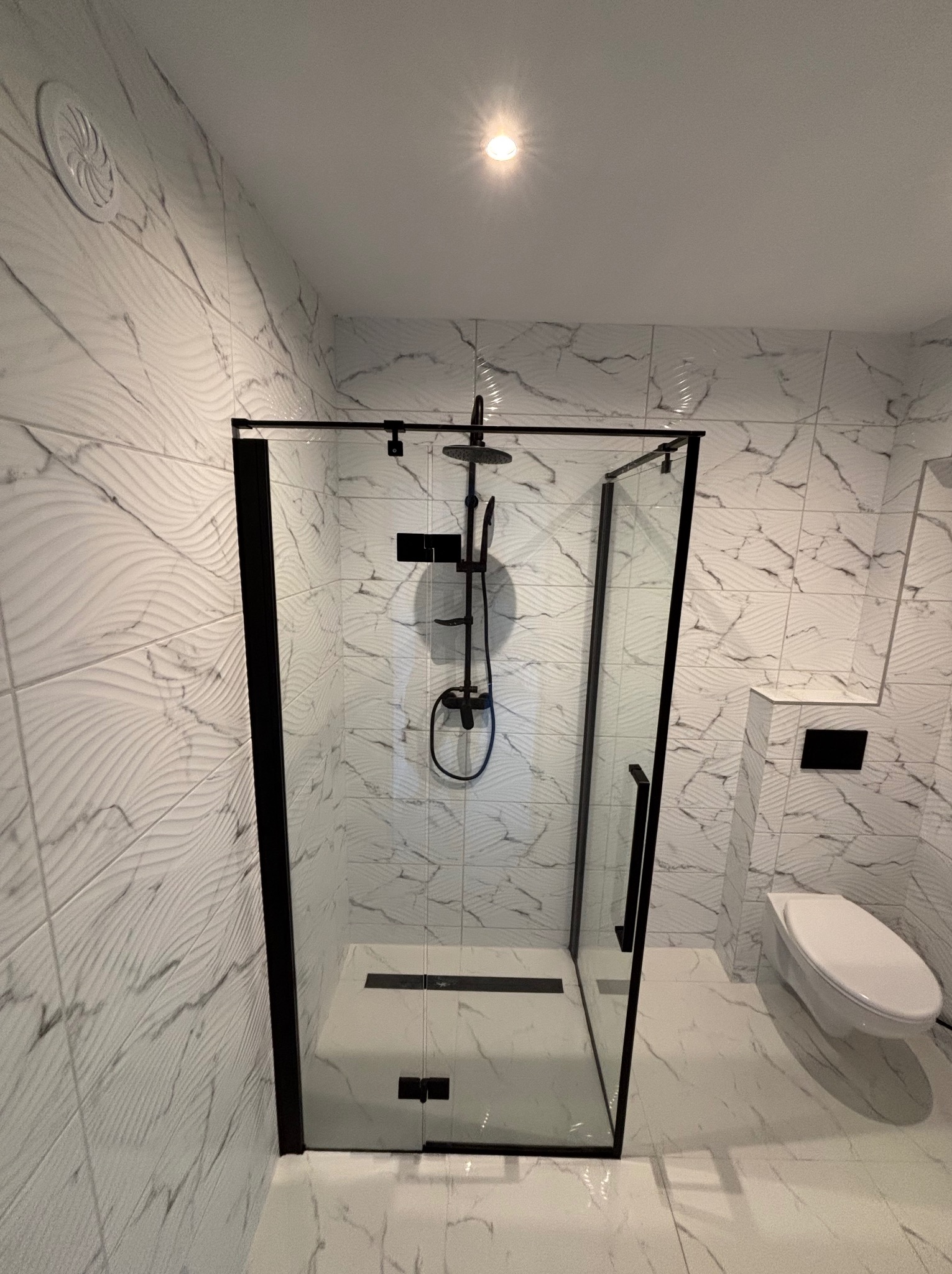 Nowoczesna kabina prysznicowa z czarnymi profilami i deszczownicą, białe płytki imitujące marmur na ścianach i podłodze, wisząca toaleta w tle. Minimalistyczny design.