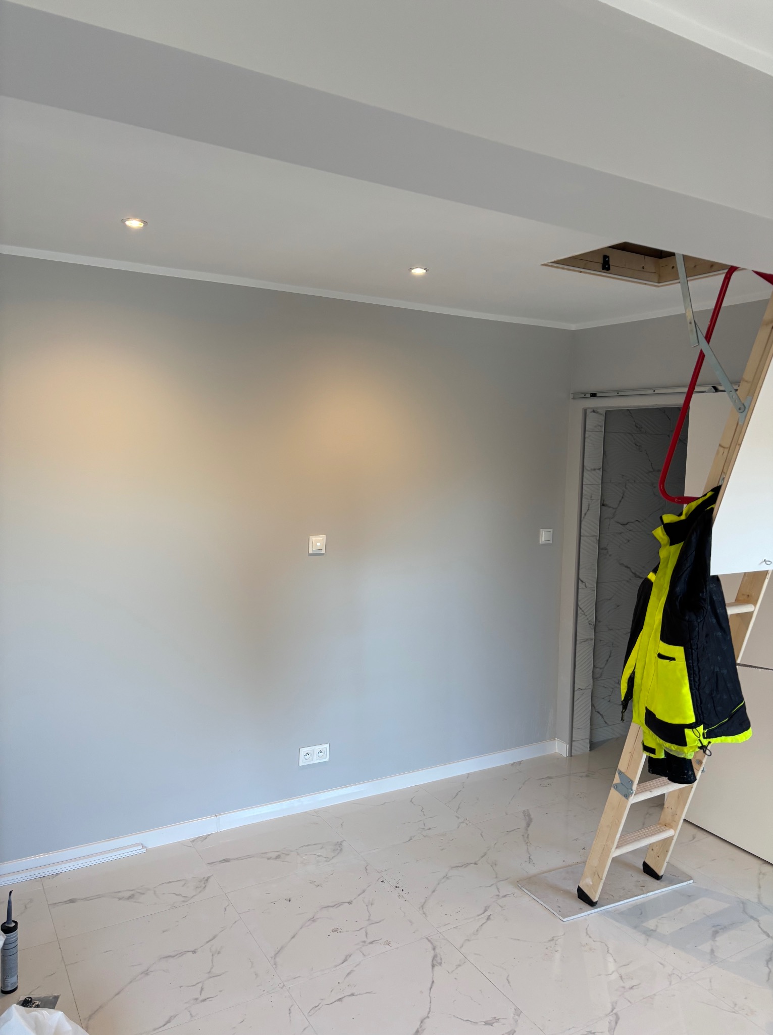 Świeżo pomalowane szare wnętrze z marmurową podłogą, widoczne gniazdka, włącznik, drabina oparta o ścianę i otwór w suficie.