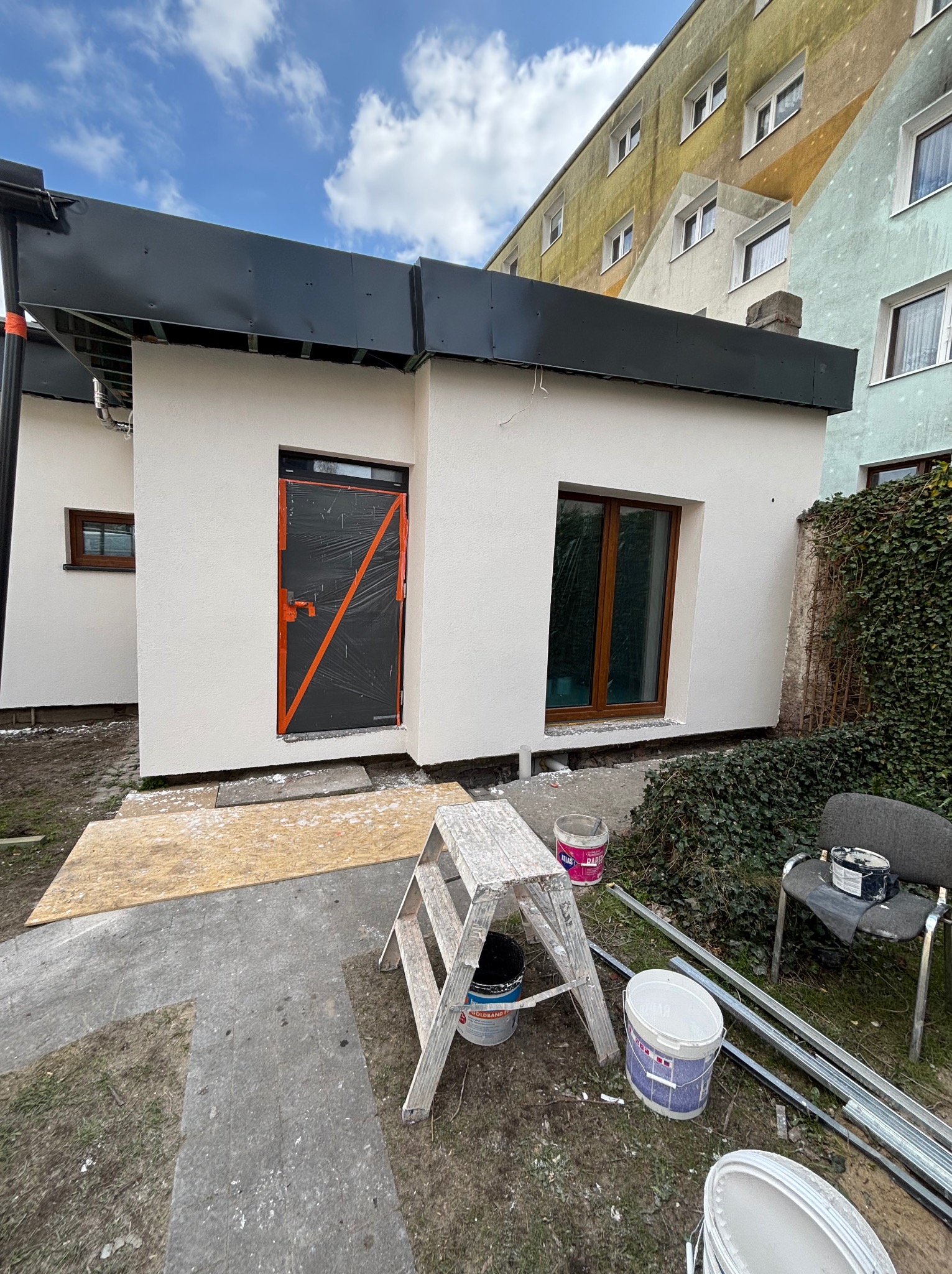 Otynkowany budynek z oknami i zabezpieczonymi drzwiami, widoczne puszki z farbą i drabina malarska. Elewacja w jasnym odcieniu, tło stanowi budynek mieszkalny.