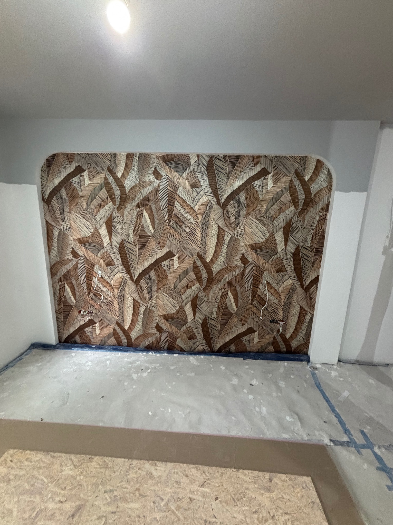 Wnęka z dekoracyjną tapetą w liście, widoczne kable elektryczne. Przygotowane podłoże pod podłogę. Szare ściany i sufit. Ujęcie z dołu, podkreślające wzór tapety.