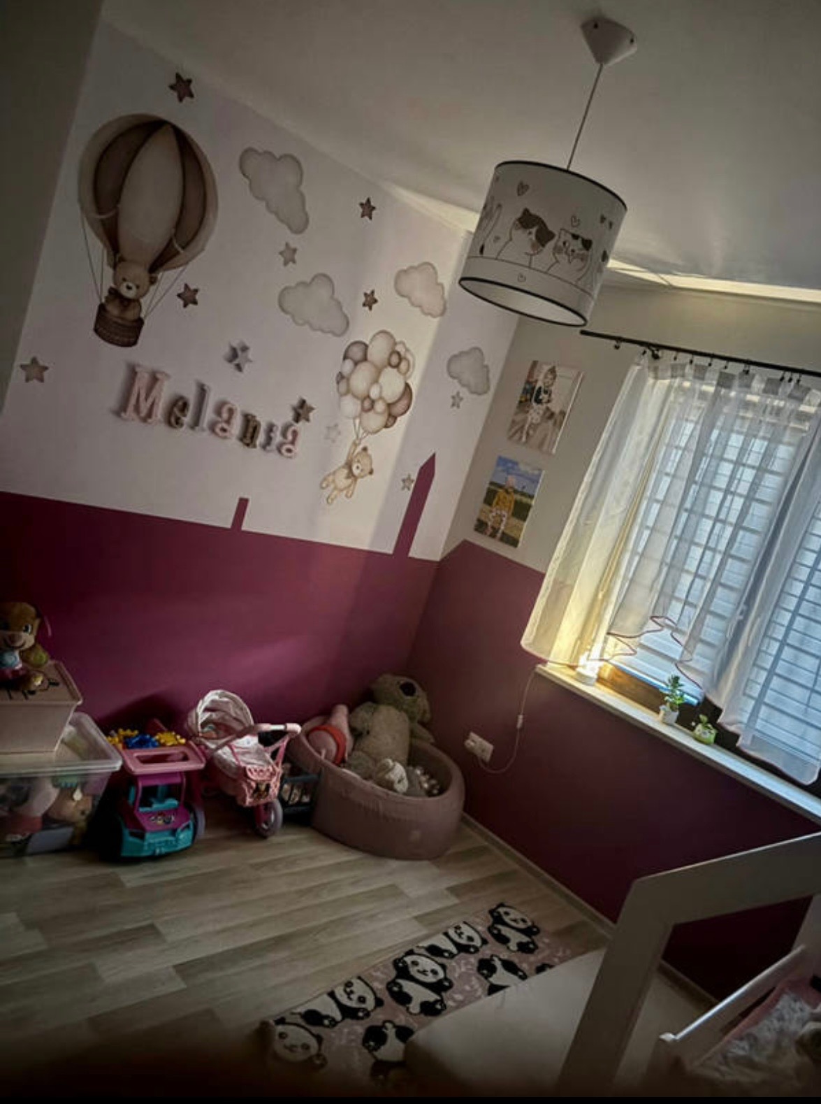 Pokój dziecięcy z dekoracyjnymi naklejkami na ścianie, lampą z motywem zwierząt i zabawkami. Ściany pomalowane w dwóch kolorach, podłoga wyłożona panelami.