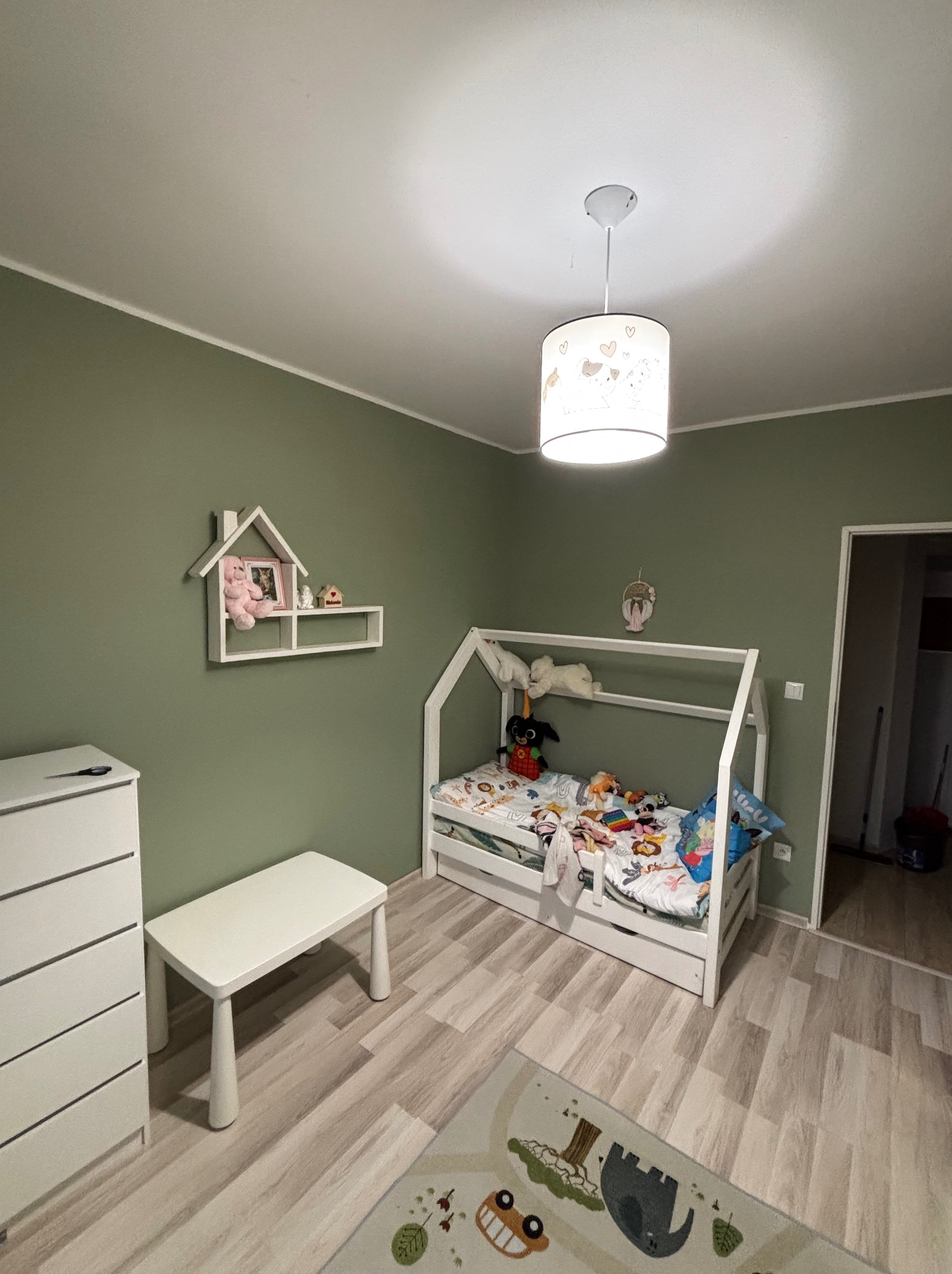 Pokój dziecięcy z łóżkiem w kształcie domku, komodą i stolikiem. Ściany w odcieniach zieleni, drewniana podłoga. Na ścianie półka w kształcie domku.