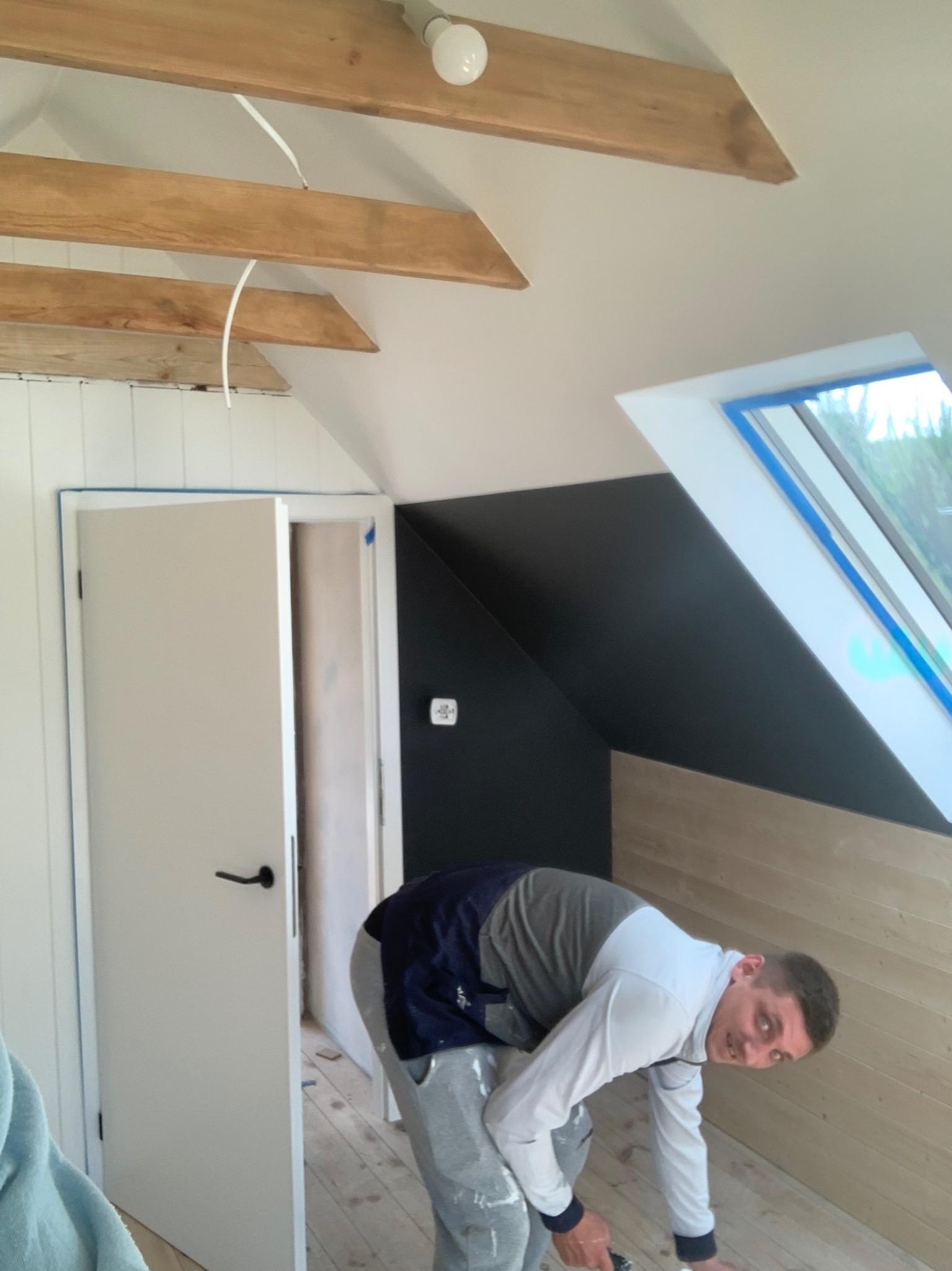 Prace wykończeniowe na poddaszu: mężczyzna szlifuje drewnianą podłogę w pomieszczeniu ze skosami, widoczne drewniane belki i okno dachowe.