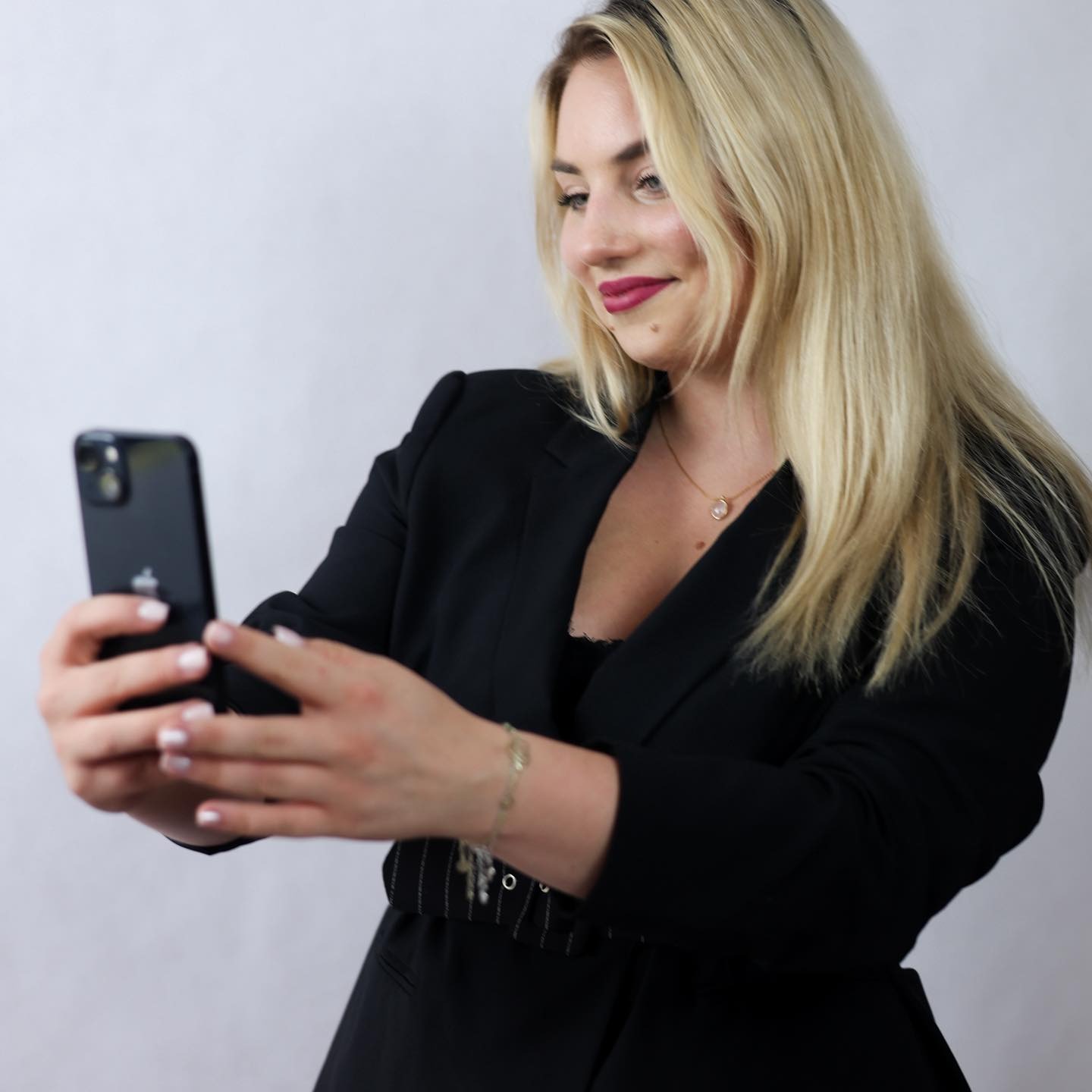 Blondynka w czarnym żakiecie robi selfie telefonem. Uśmiechnięta kobieta z biżuterią pozuje na jasnym tle, trzymając smartfon w dłoniach.