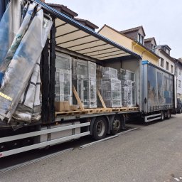 Thermo-plast Sp.z o.o. - Ciężarówka z naczepą, częściowo odsłonięta plandeka, załadowana oknami PCV w folii, na paletach. Widok z boku na tle zabudowy miejskiej.