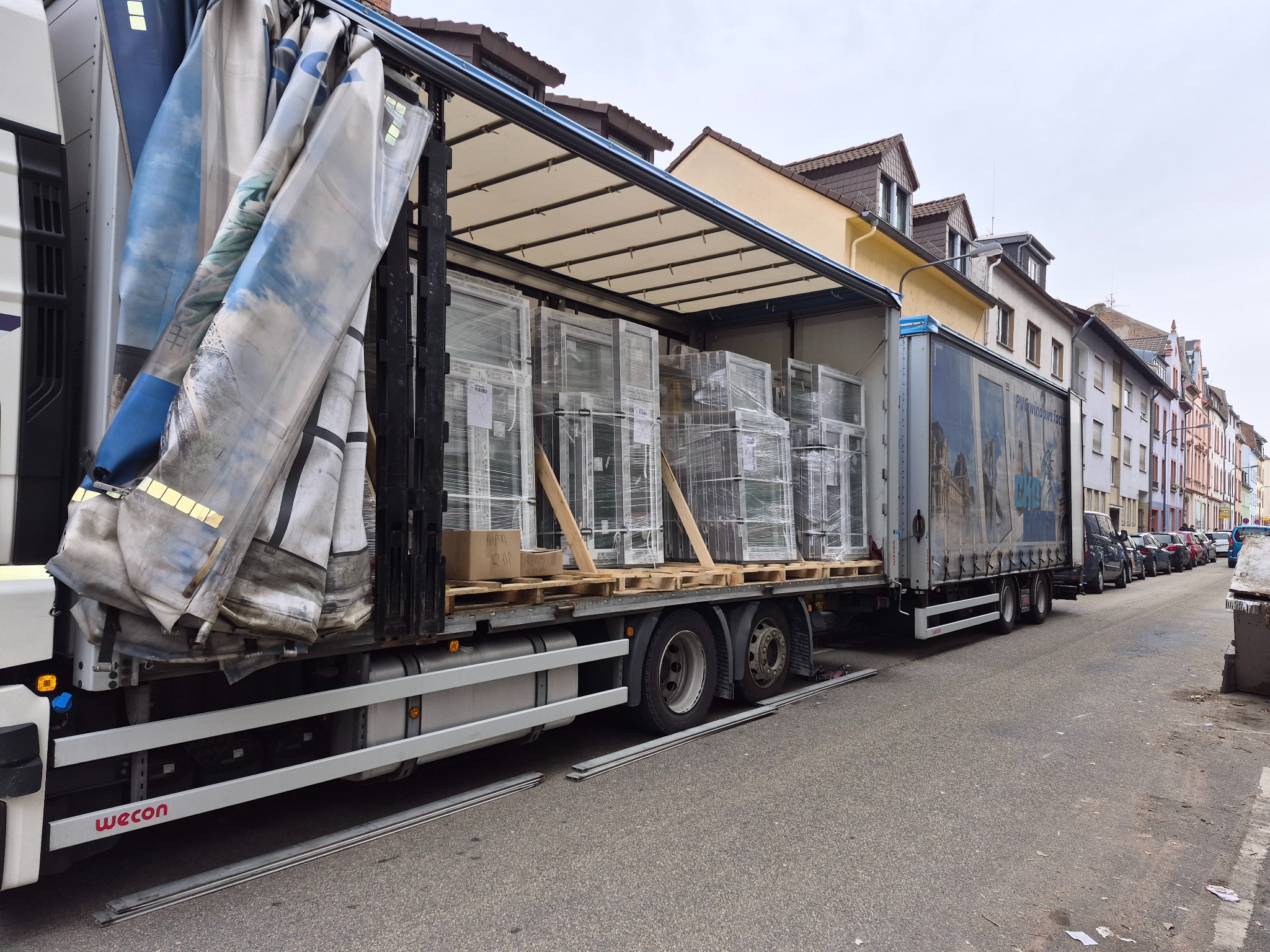 Ciężarówka z naczepą, częściowo odsłonięta plandeka, załadowana oknami PCV w folii, na paletach. Widok z boku na tle zabudowy miejskiej.