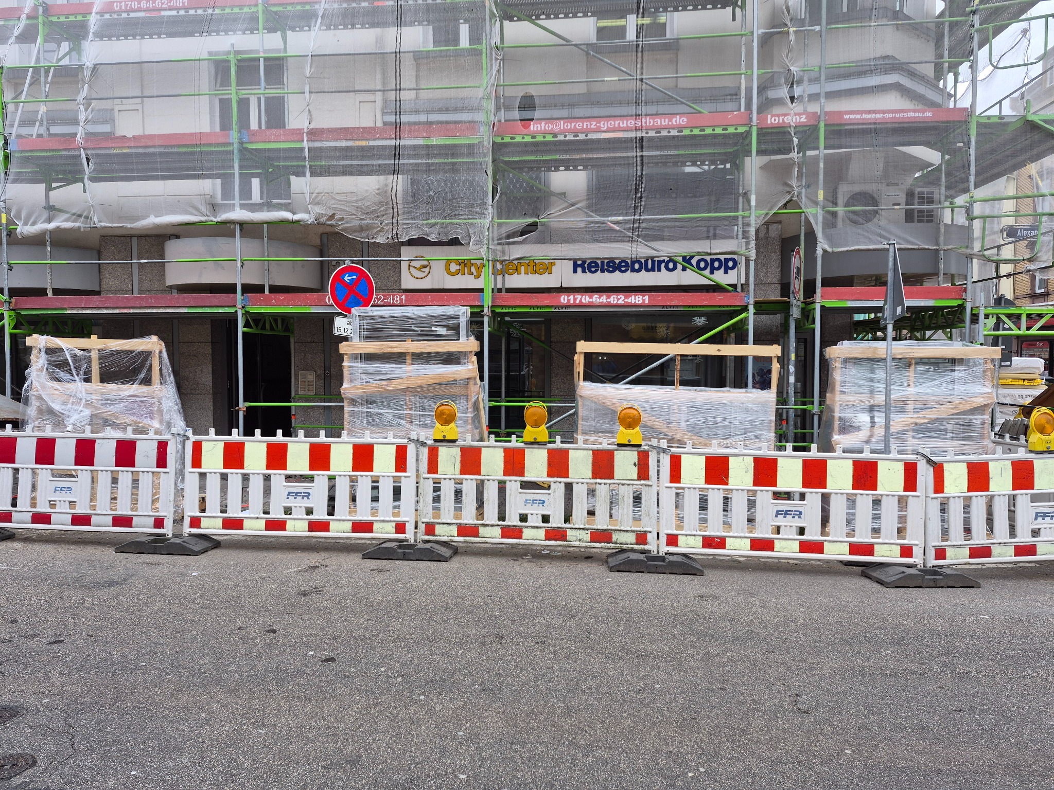 Budynek w rusztowaniach, zabezpieczony barierami z żółtymi lampami ostrzegawczymi. Widoczne okna i drzwi w folii ochronnej. Prace remontowe w toku.
