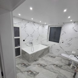 Wykończona łazienka z wanną obudowaną płytkami imitującymi marmur, białą armaturą i oświetleniem punktowym. Płytki ścienne i podłogowe w tym samym wzorze.