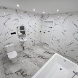 Wykończona łazienka z marmurowymi płytkami na ścianach i podłodze. Widoczna toaleta, umywalka, wanna i białe drzwi z czarnymi akcentami. Nowoczesny design.