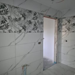 Wykończona łazienka z białymi płytkami imitującymi marmur i pasem dekoracyjnym z motywem liści. Widoczne otwory na armaturę i fragment ościeżnicy drzwiowej.