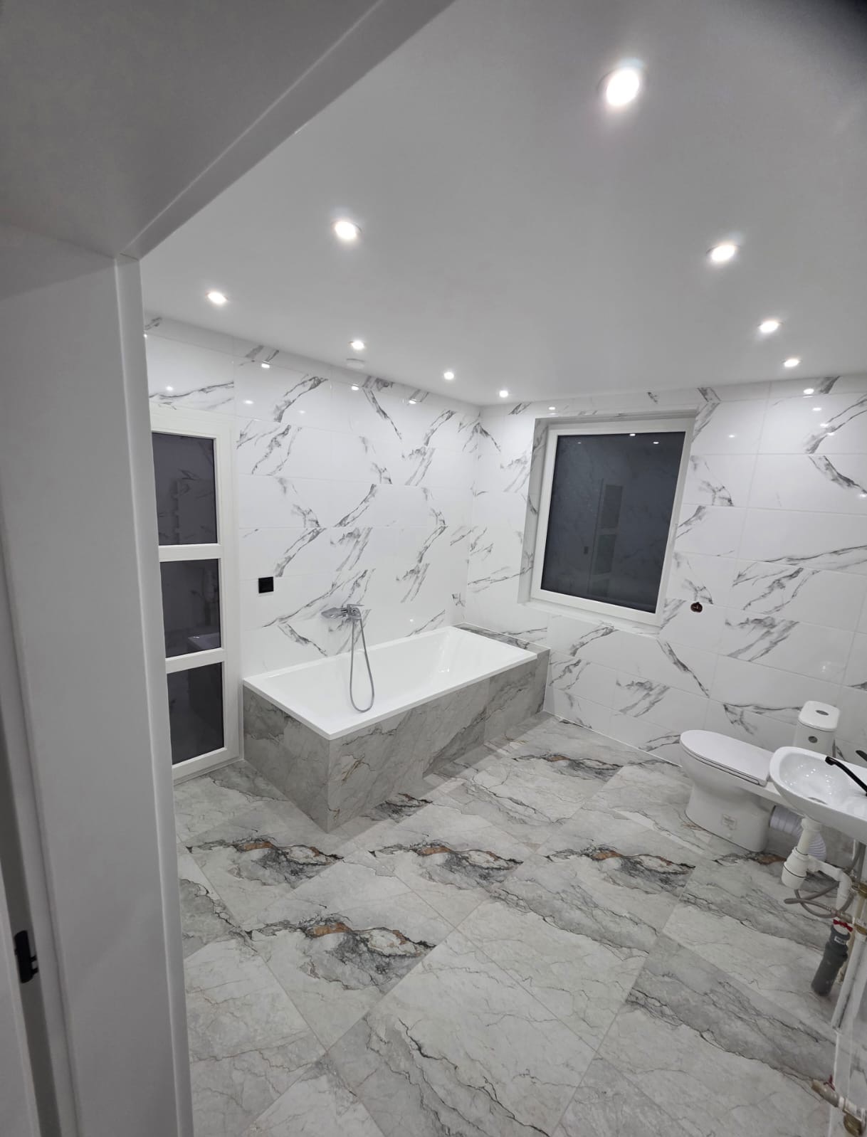 Wykończona łazienka z wanną obudowaną płytkami imitującymi marmur, białą armaturą i oświetleniem punktowym. Płytki ścienne i podłogowe w tym samym wzorze.