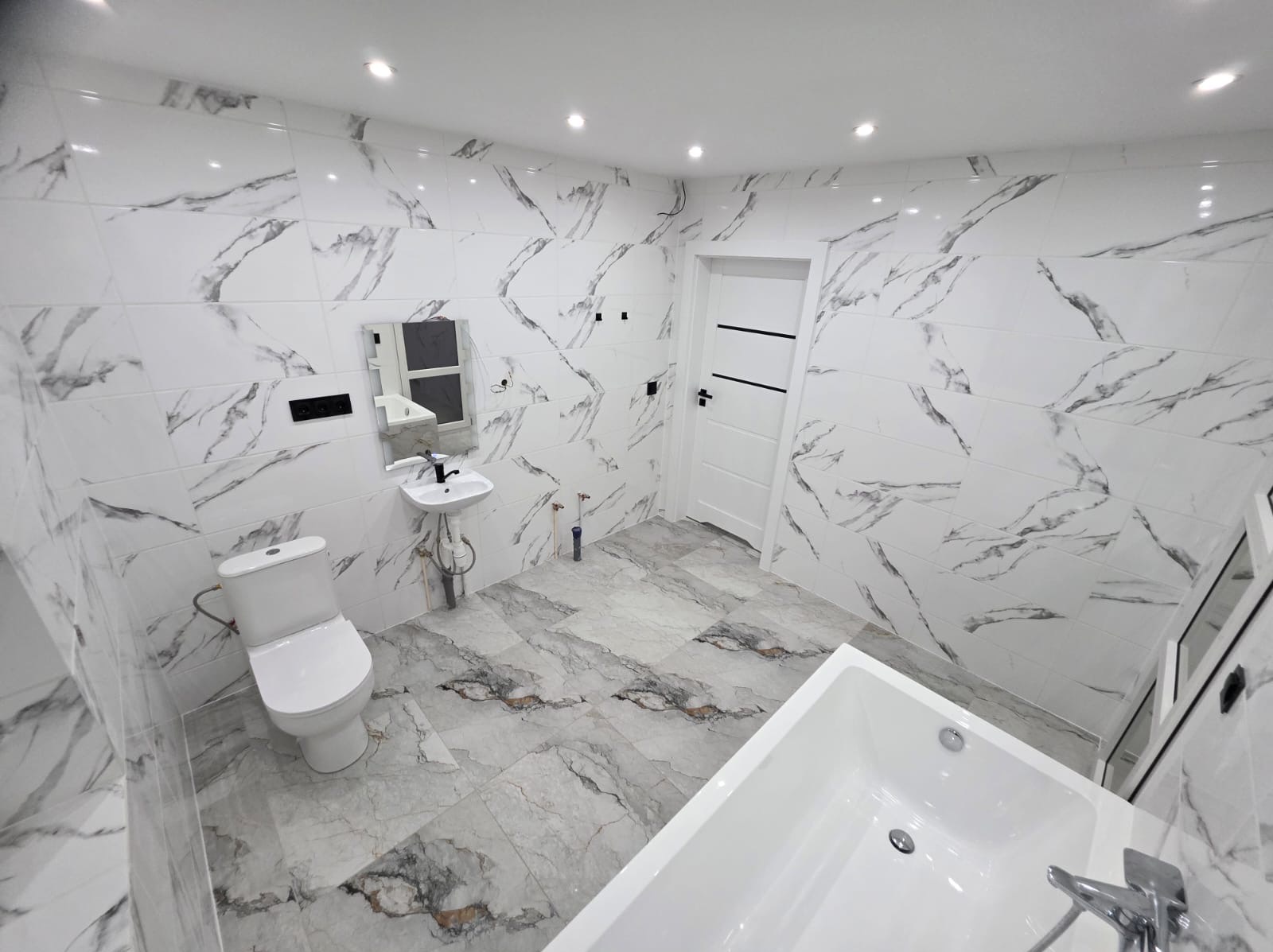 Wykończona łazienka z marmurowymi płytkami na ścianach i podłodze. Widoczna toaleta, umywalka, wanna i białe drzwi z czarnymi akcentami. Nowoczesny design.