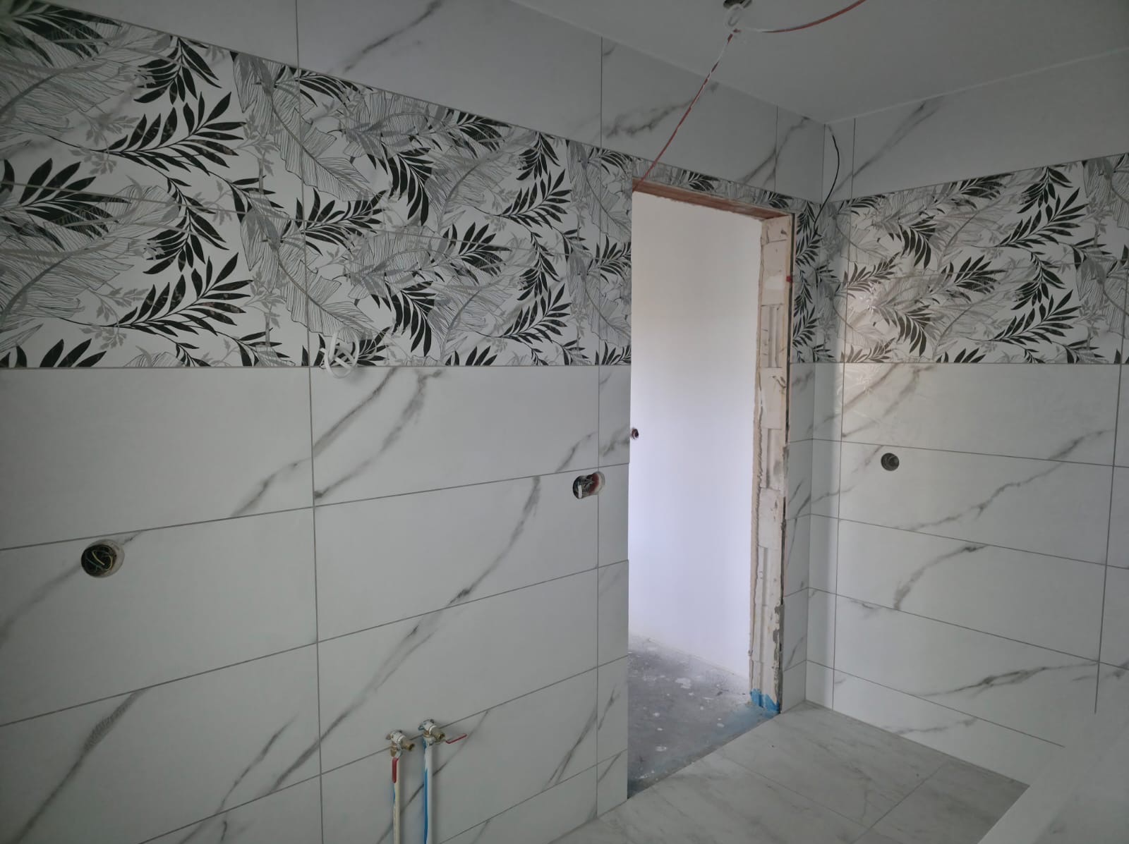 Wykończona łazienka z białymi płytkami imitującymi marmur i pasem dekoracyjnym z motywem liści. Widoczne otwory na armaturę i fragment ościeżnicy drzwiowej.