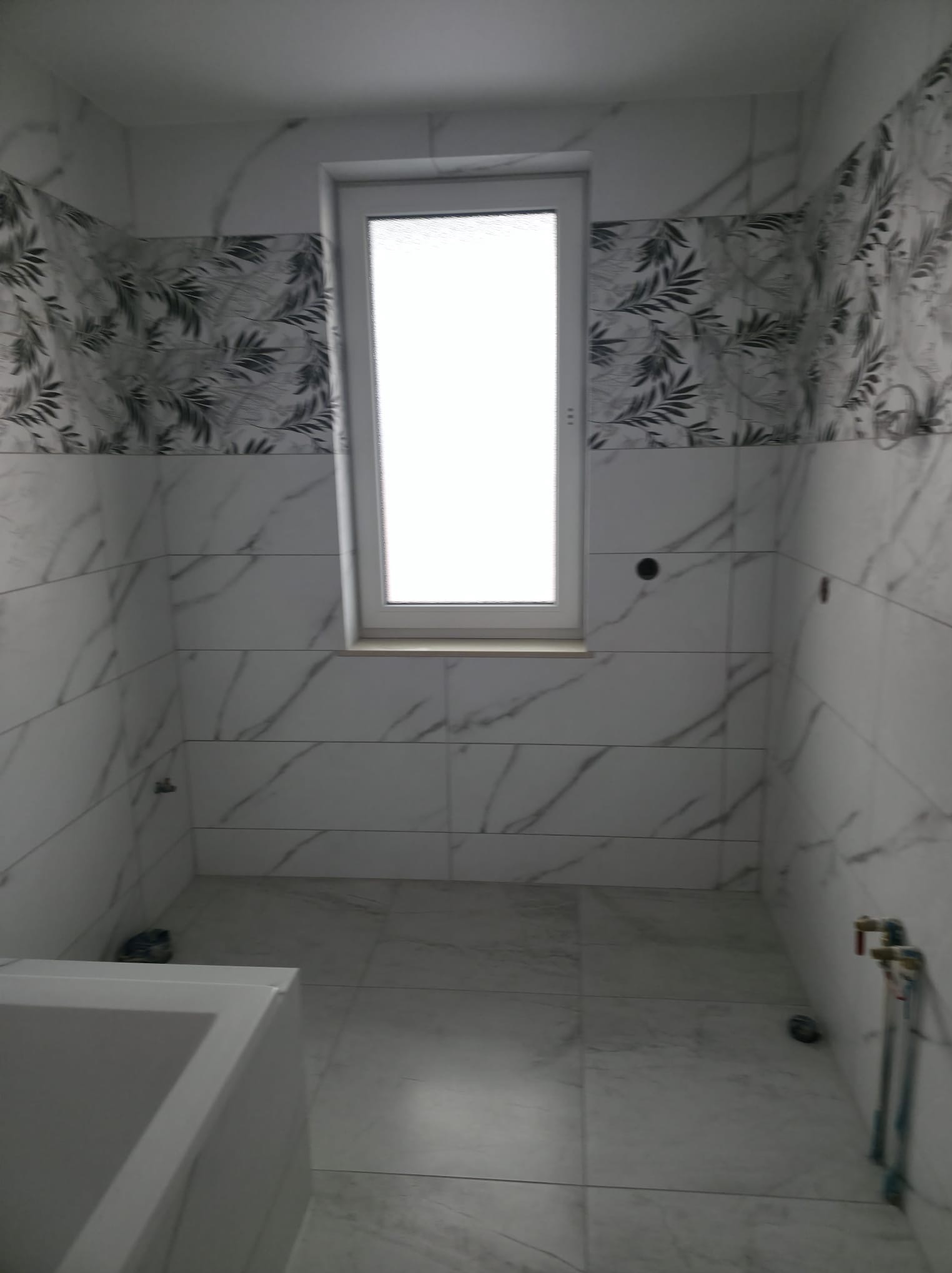 Wykończona łazienka z białymi płytkami imitującymi marmur i dekoracyjnym pasem z motywem liści. Widoczne okno i fragment wanny. Minimalistyczny design.