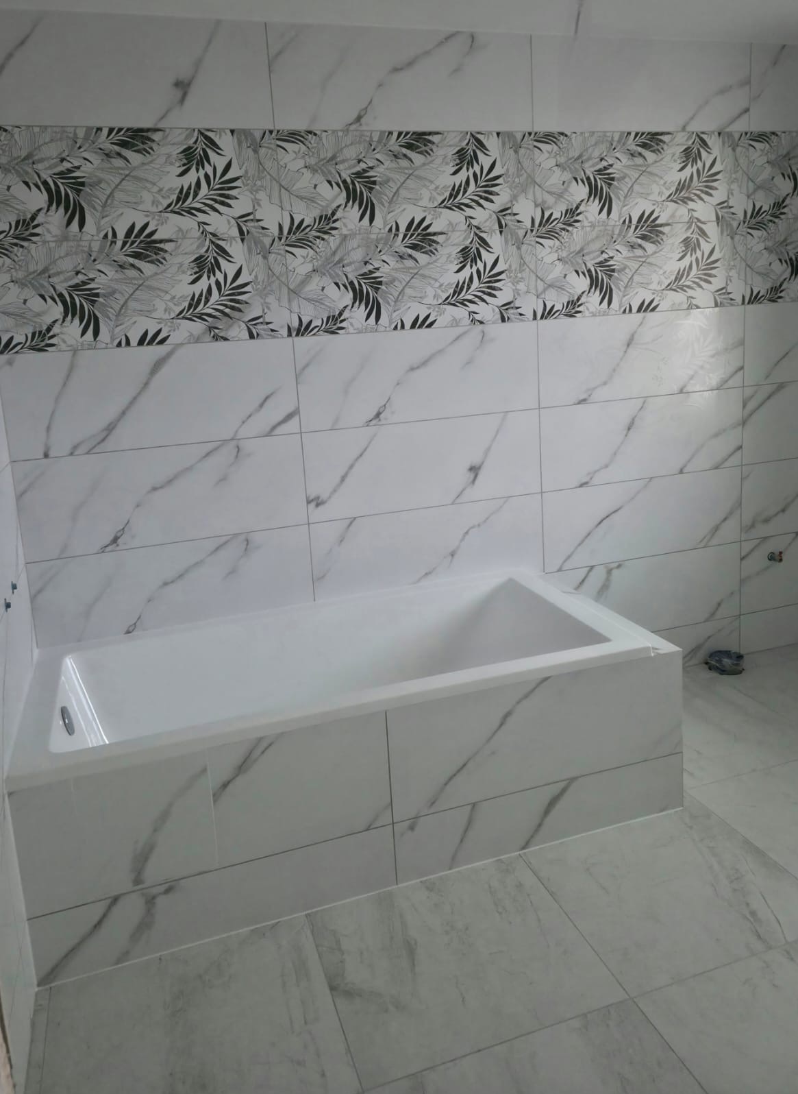 Wykończona łazienka z wanną obudowaną płytkami imitującymi marmur. Nad wanną pas płytek z motywem roślinnym. Podłoga wyłożona szarymi płytkami.