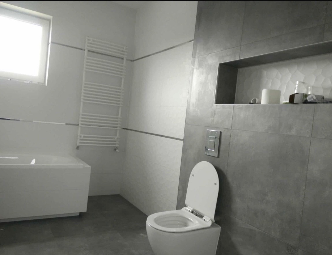 Nowoczesna łazienka z szarymi płytkami, białą armaturą i grzejnikiem drabinkowym. Wnęka w ścianie z dekoracyjną fakturą i akcesoriami. Minimalistyczny design.